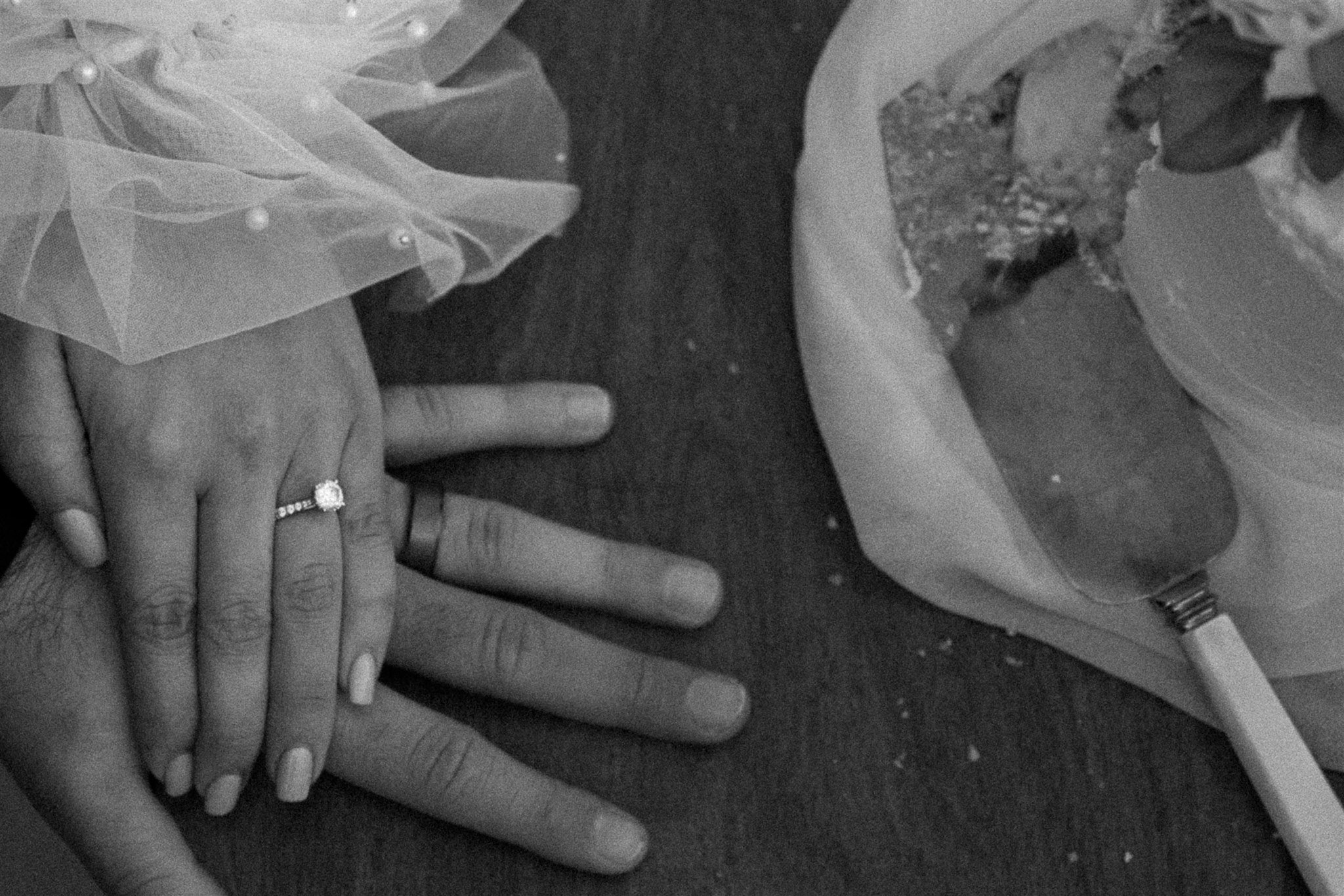 Couple is at a table with a wedding cake that has been cut into and there is a wedding cake knife lying there. The couple isn't shown but their hands rest on top of each other to show their wedding bands