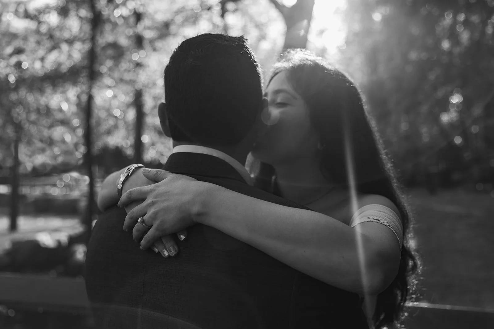 Couple poses for a photo while one partner kisses the other one on the cheek. The image is in black and white but a sun ray is lighting their heads making the image look whimsical. You can see their faces but you can tell they are married with rings