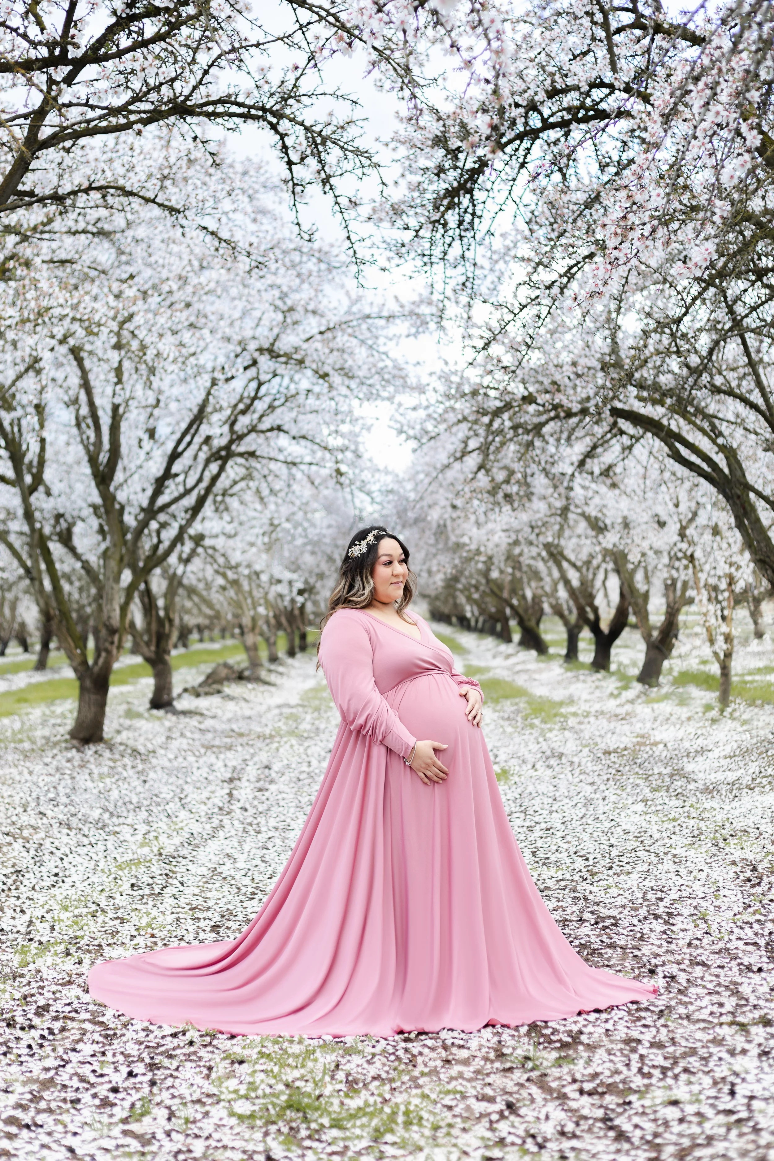 A pregnant mom stands in the middle of a path surrounded by almond blossoms in the Central Valley California. She wears a pink gown that falls to the floor as she holds her belly and looks forward with a soft and calm expression
