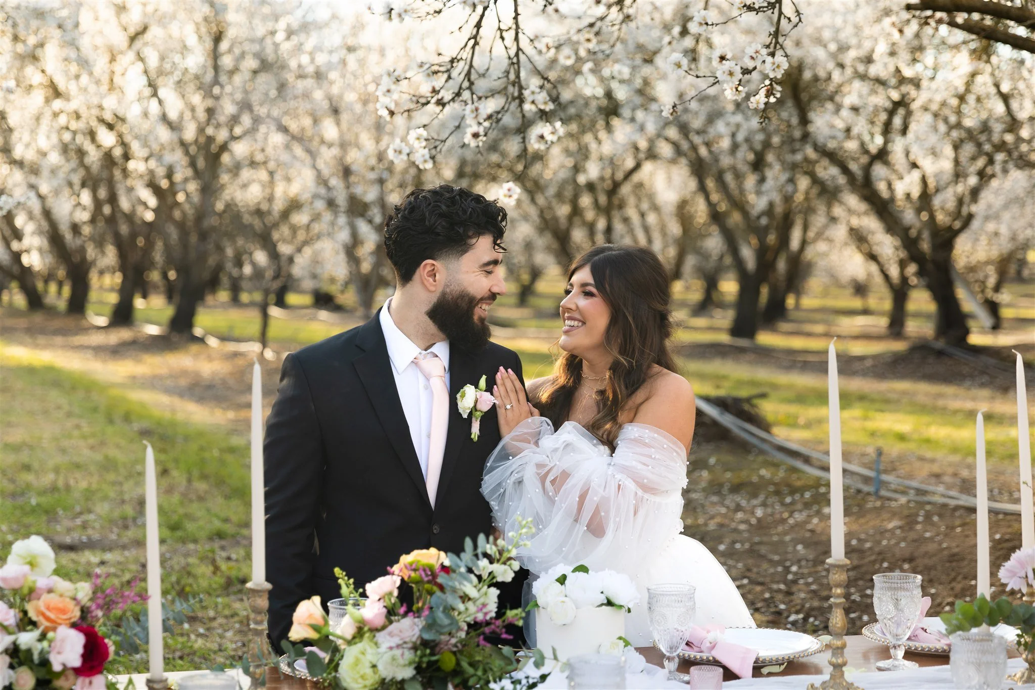 Married couple sit with each other at luxury picnic table and surrounded by almond blossoms in Merced California
