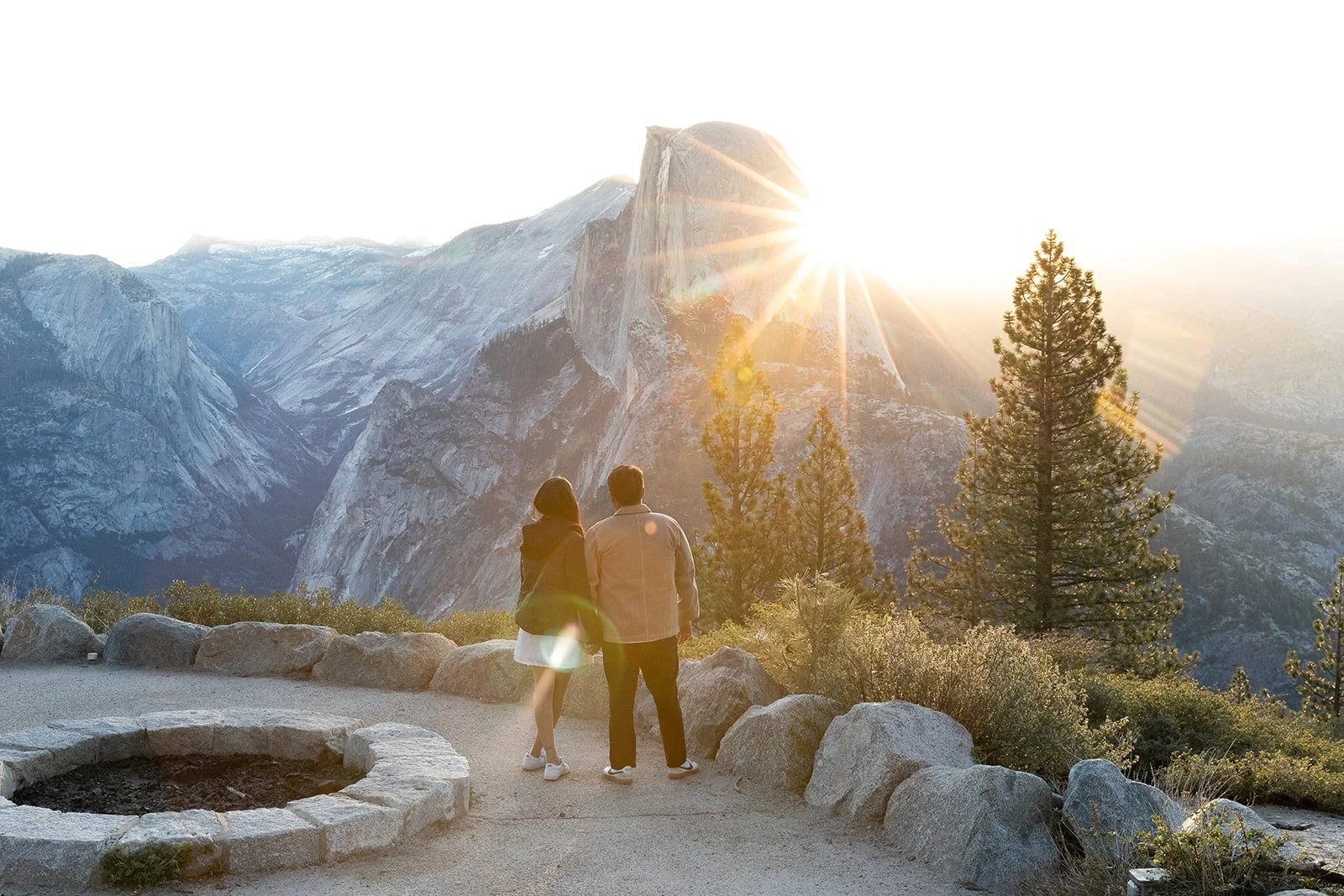 couple stands on paved trail overlooking Yosemite Valley  as the sun peeks out of Half Dome after sunrise