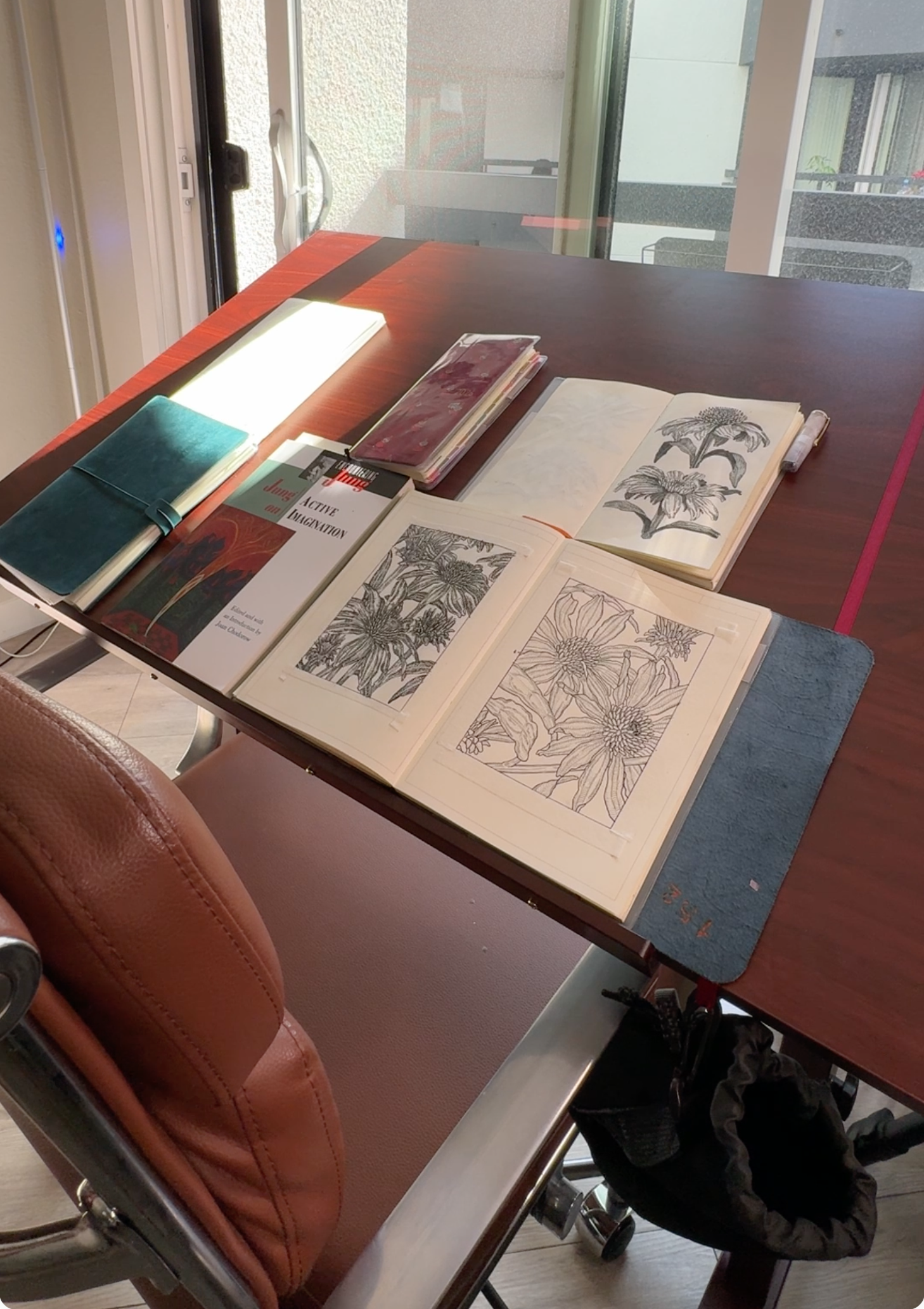 Table with open botanical illustration books and notebooks, set in a room with glass doors and balcony.
