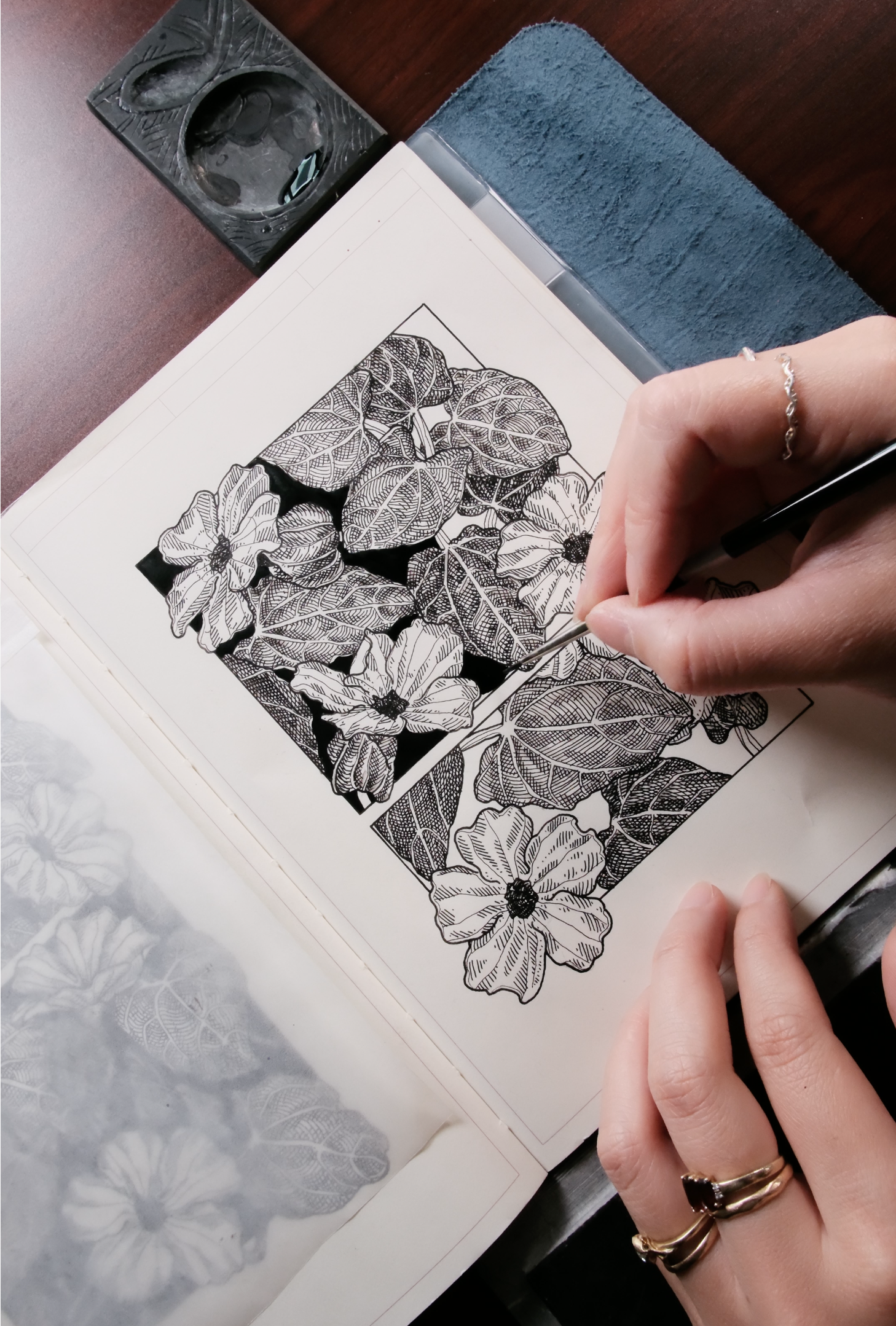 A person is creating detailed black and white floral artwork in a sketchbook, using a fine-tipped pen to illustrate large flowers and leaves with intricate line work.