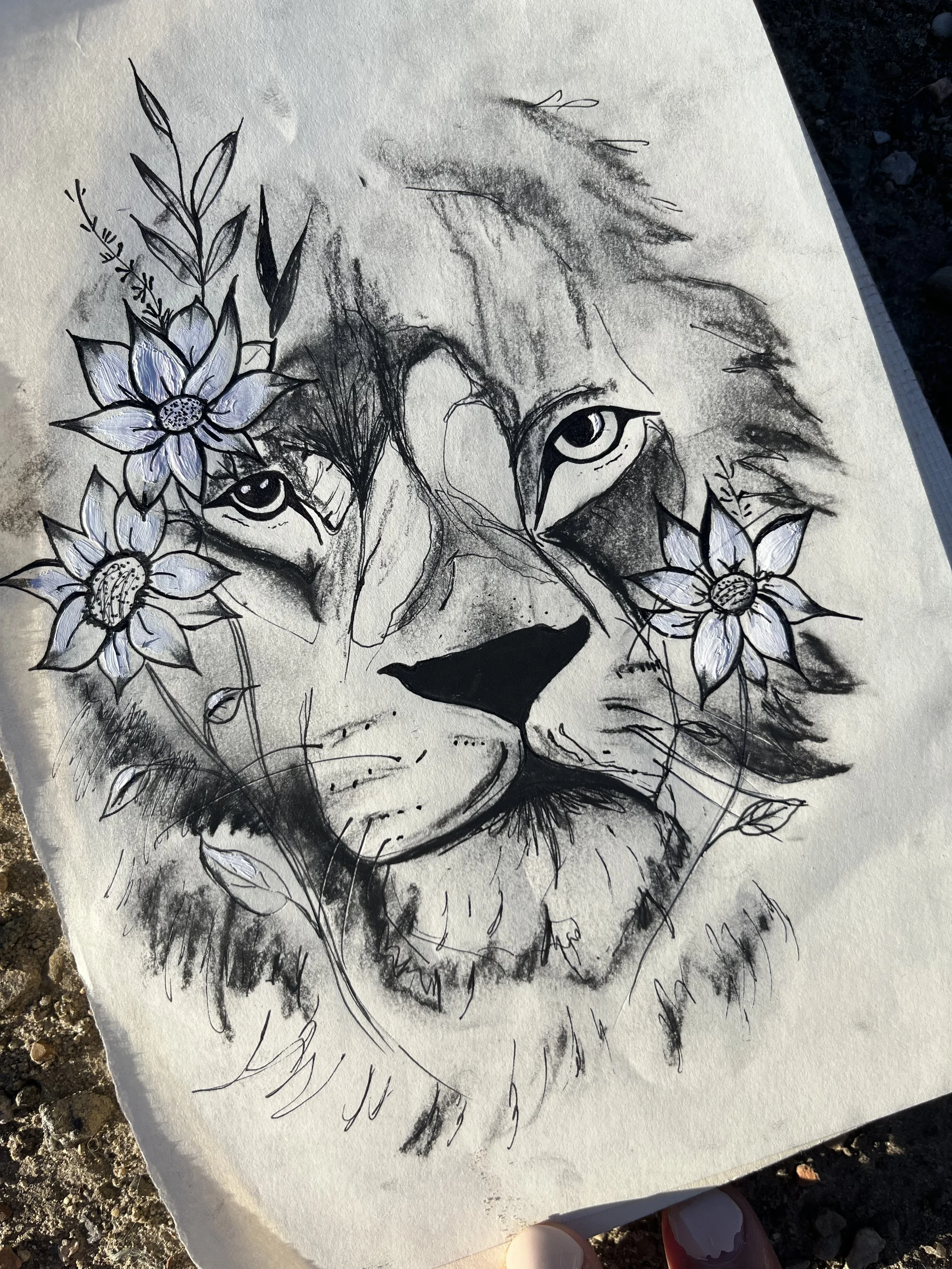 A black and white sketch of a lion's face with large eyes and detailed mane, surrounded by hand-drawn flowers with shaded petals.