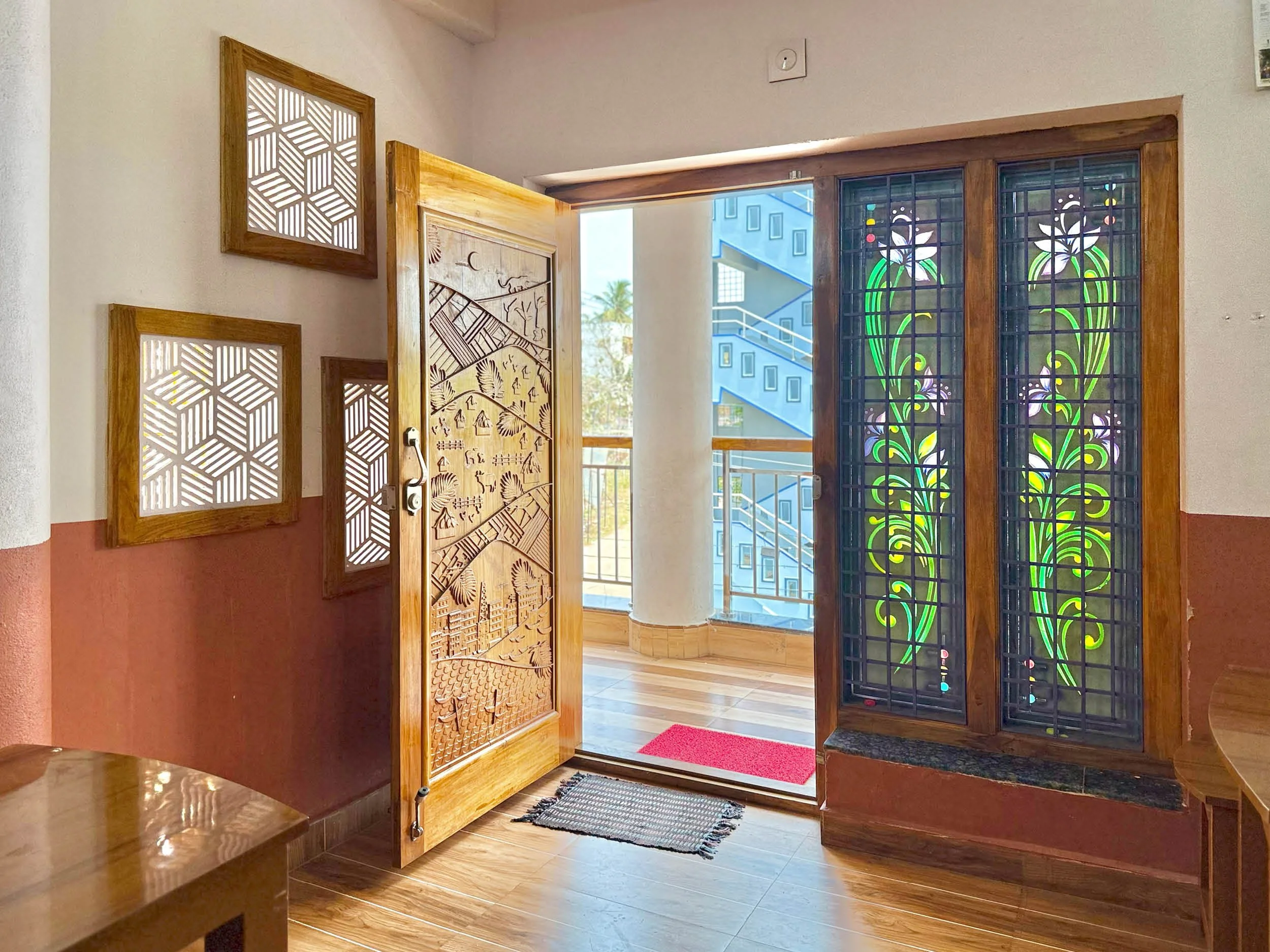 An interior view of a doorway with decorative stained glass windows, a carved wooden door, and wooden accents. There is a red welcome mat and a rug in front of the door, with stairs visible outside.