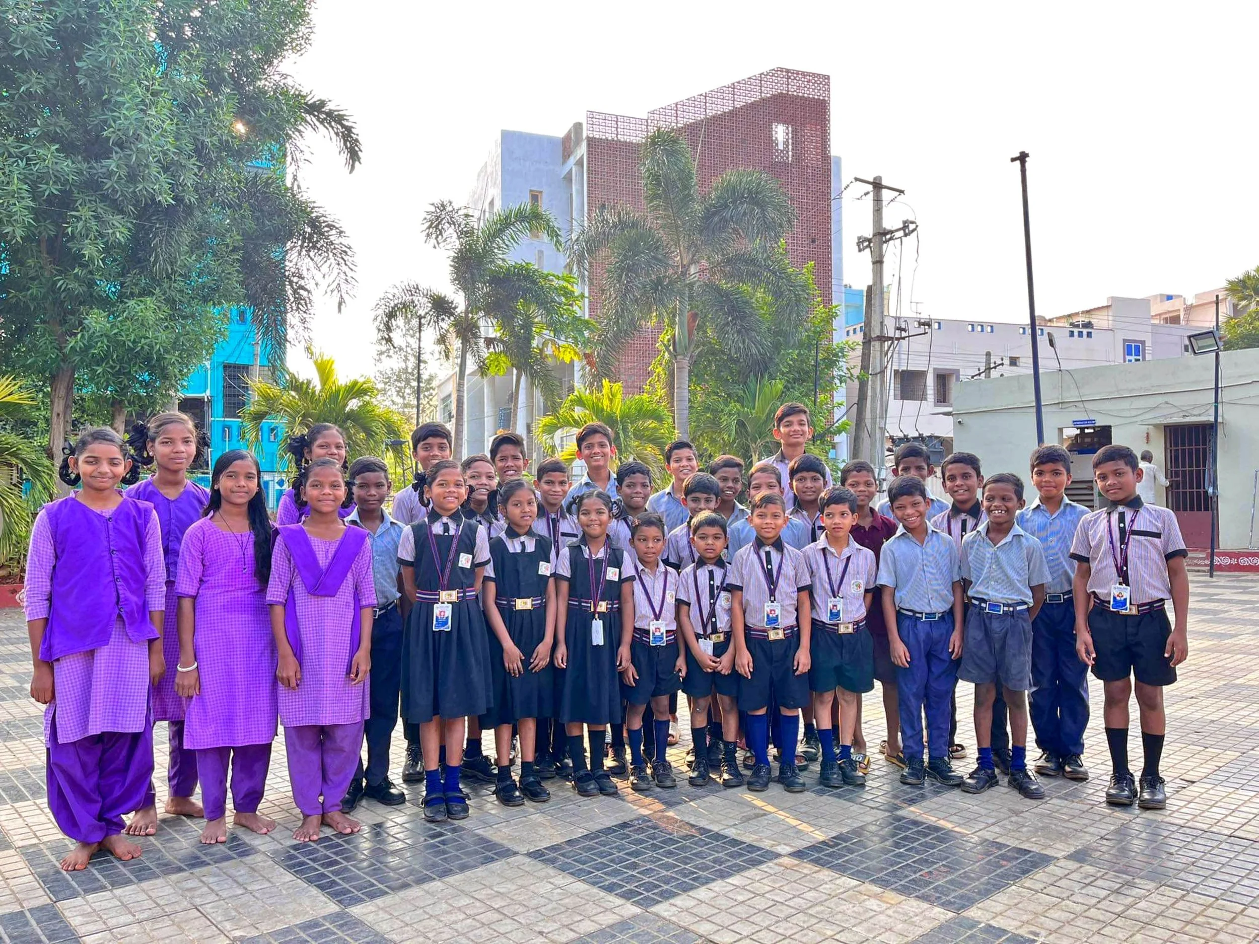 Group of school children posing outdoors, with school buildings, trees, and utility poles in the background, during daytime.