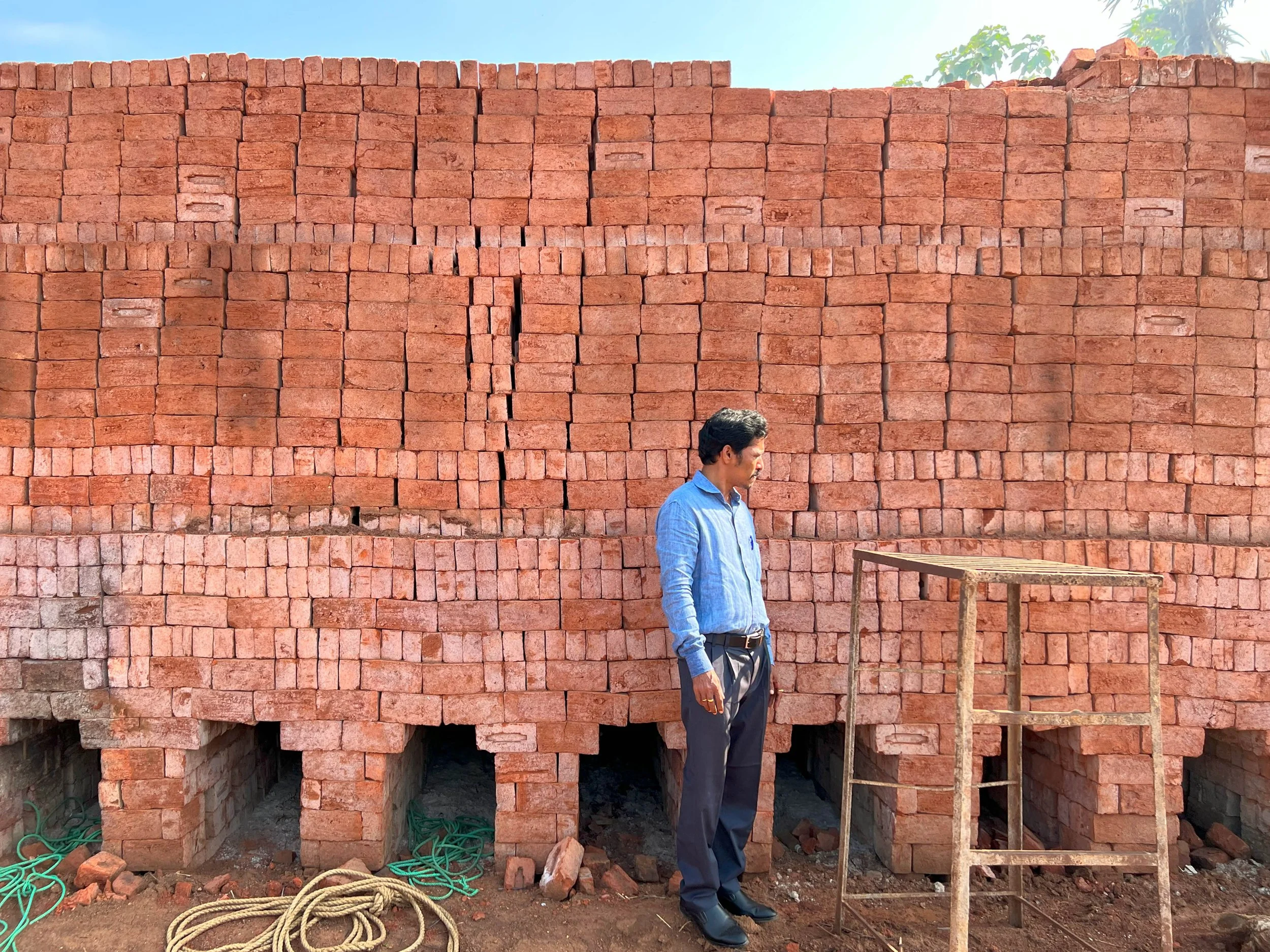 A man stands in front of a large brick wall that is under construction. The wall is made of red bricks stacked neatly. The man is wearing a light blue shirt and dark pants, looking to his left. Construction materials, including wooden scaffolding and