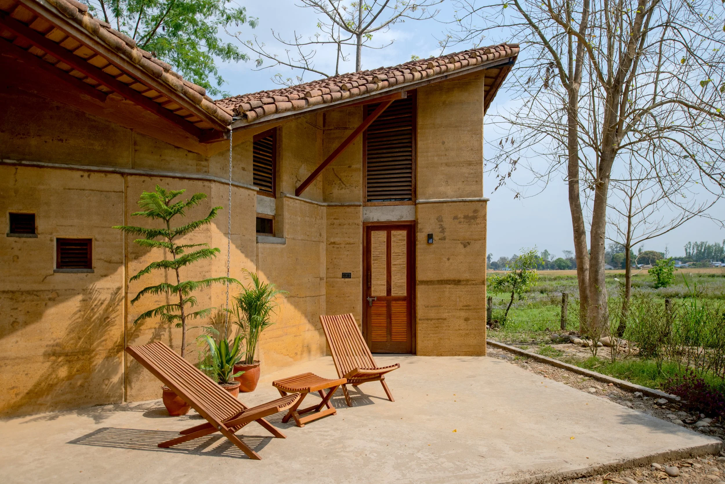 A house with a clay-tiled roof, beige walls, and wooden window shutters. In front of the house, there are three wooden lounge chairs and potted plants on a concrete patio. A large leafless tree is beside the house, and open green fields with some tre