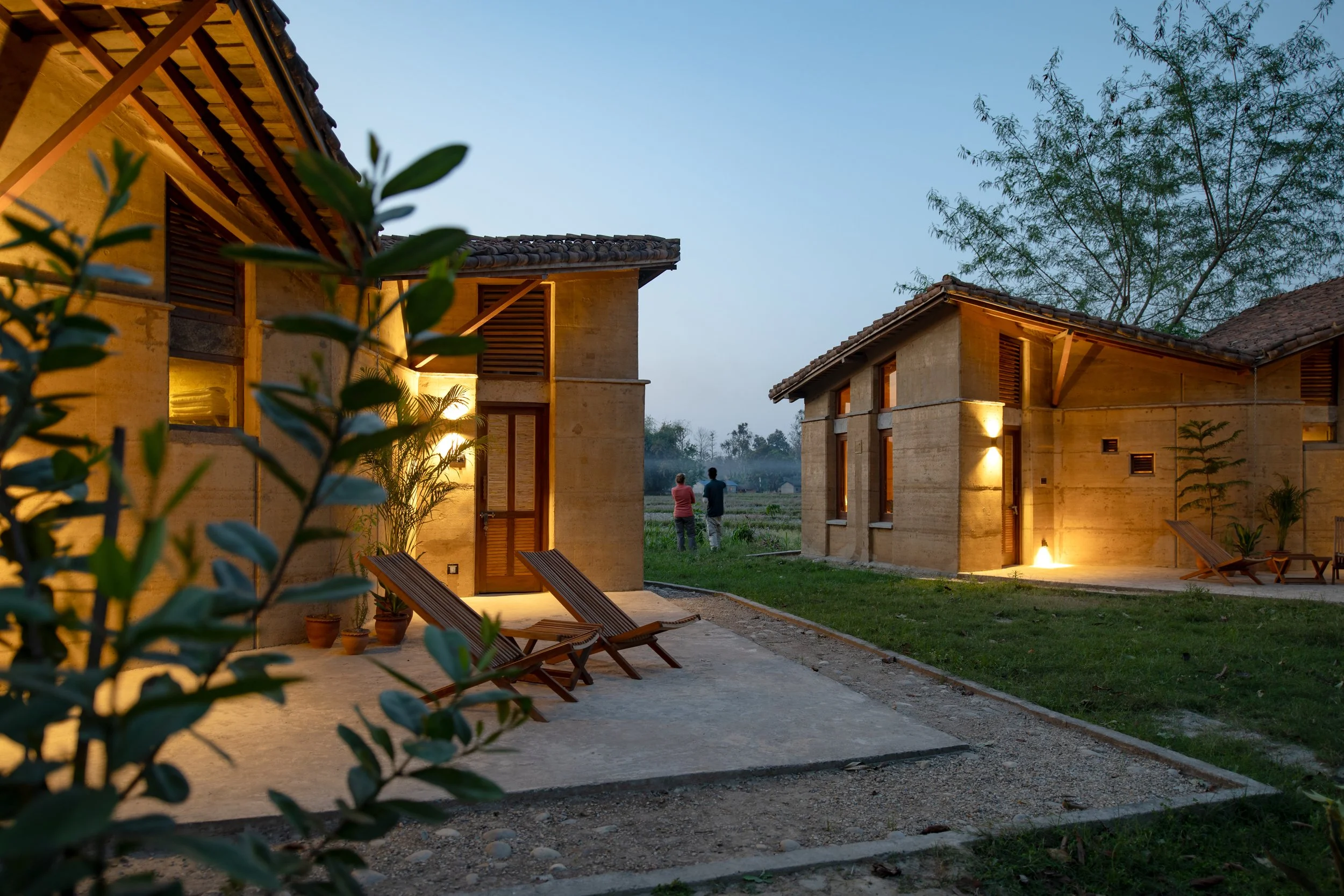 Exterior view of modern houses at twilight with warm lighting, wooden shutters, and lounge chairs in the yard, two people walking in the distance, surrounded by greenery and trees.