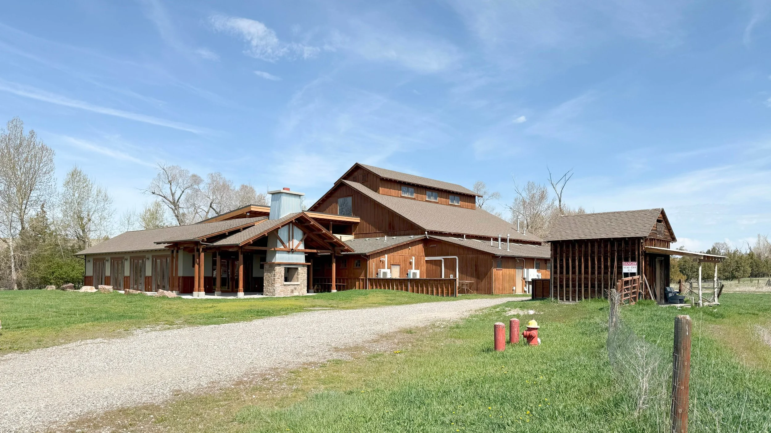 The central building of the Lighthouse Ranch in Montana with multiple roofs and windows, surrounded by grass and trees under a partly cloudy sky.