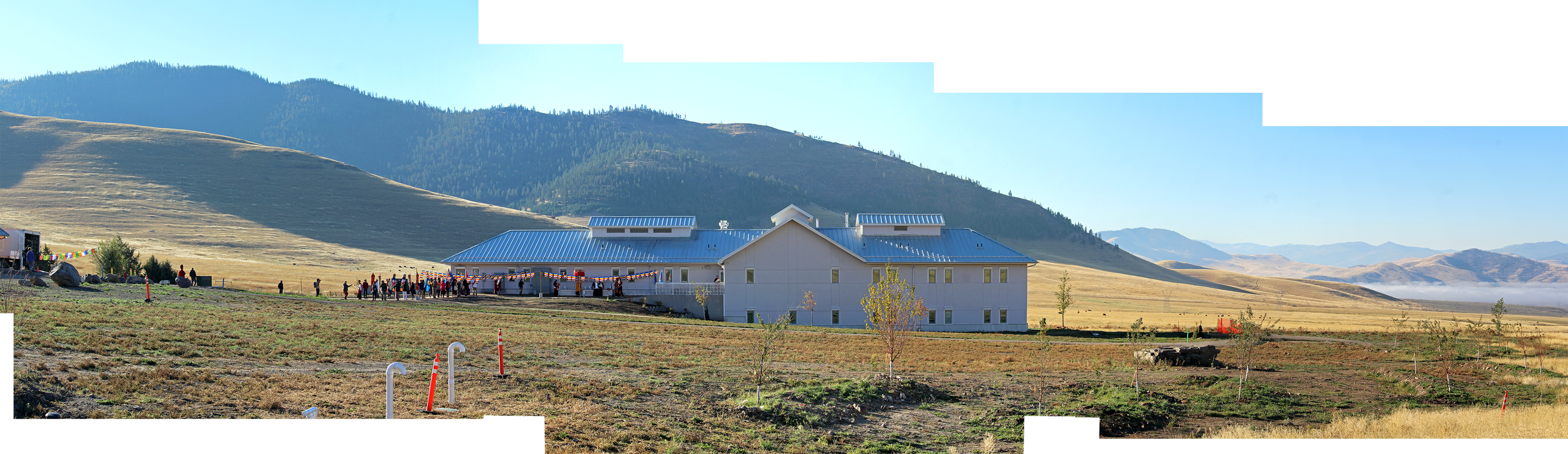 A large building with a metal roof in a mountainous area. There are small trees and a group of people gathered outside, and rolling hills in the background under a clear blue sky.