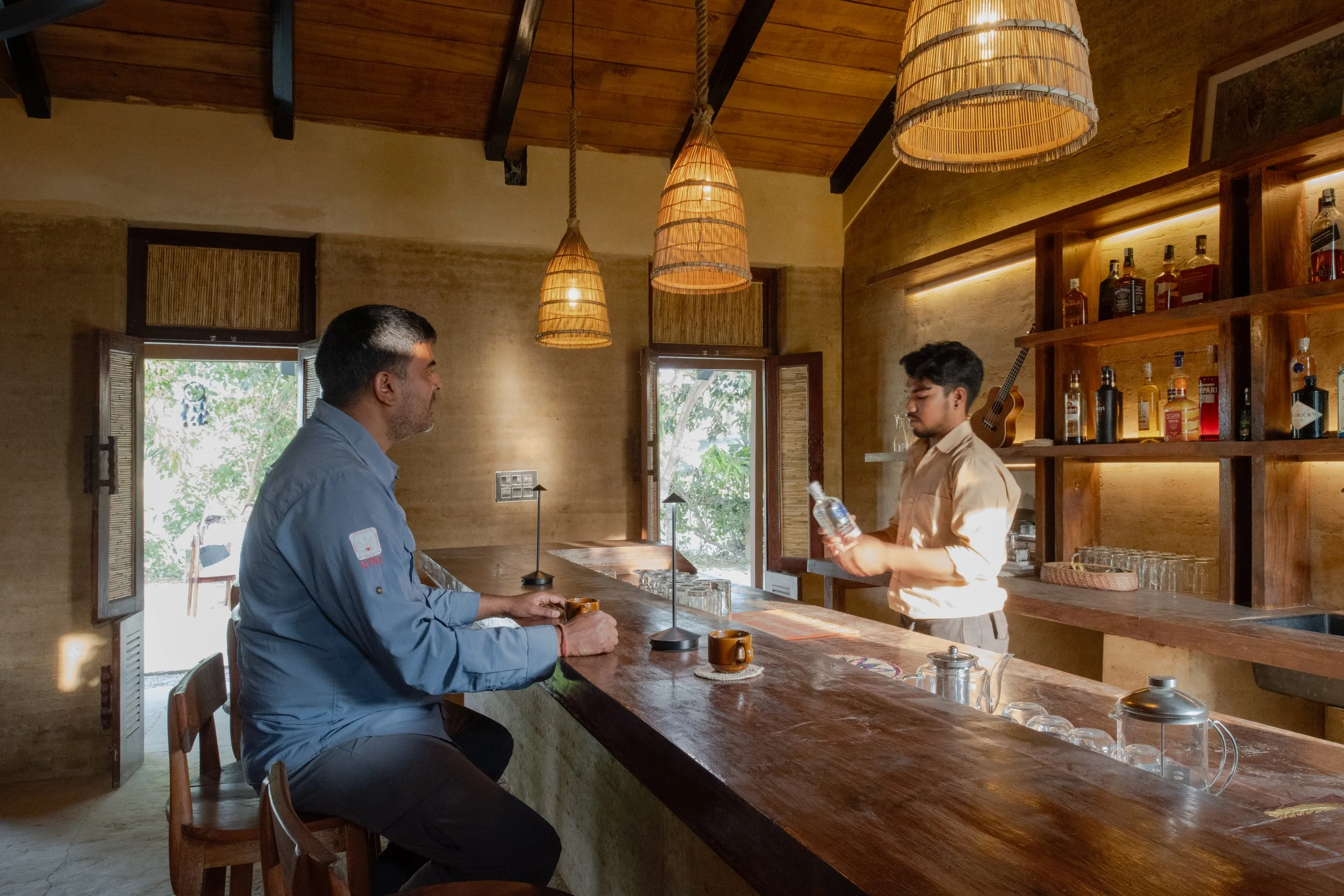 A man in a blue shirt sitting at a wooden bar counter with a bartender in a beige shirt serving drinks in a cozy, warmly lit bar with wooden shelves and bottles on display.