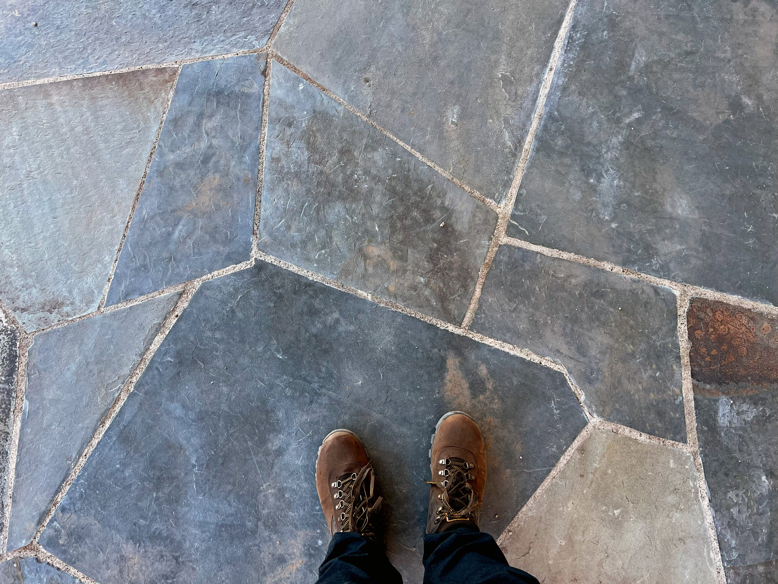 View of a person's brown boots standing on a tiled stone floor with irregular shapes and grayish-blue hues, with the edge of another shoe visible at the bottom of the image.