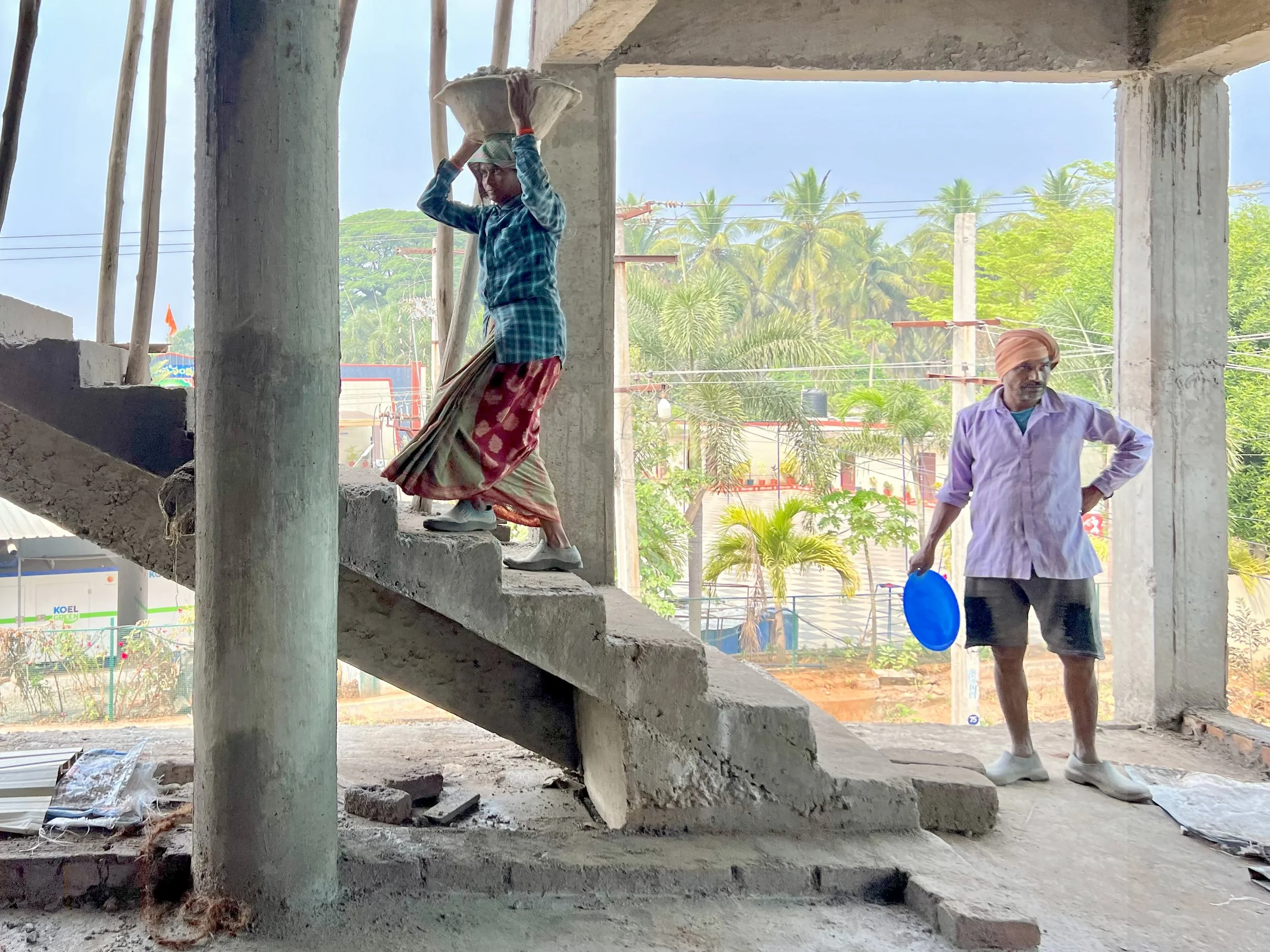 Two men are at a construction site inside a building under construction. One man is standing on a staircase and carrying a bag on his head. The other man is standing on the ground holding a blue water container. The background shows trees and power l