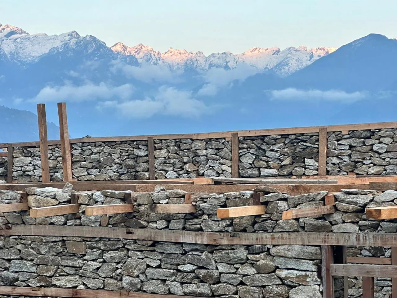 A timber-laced stone masonry wall under construction with the snow-capped peaks of the Himalayan mountain range in the background.