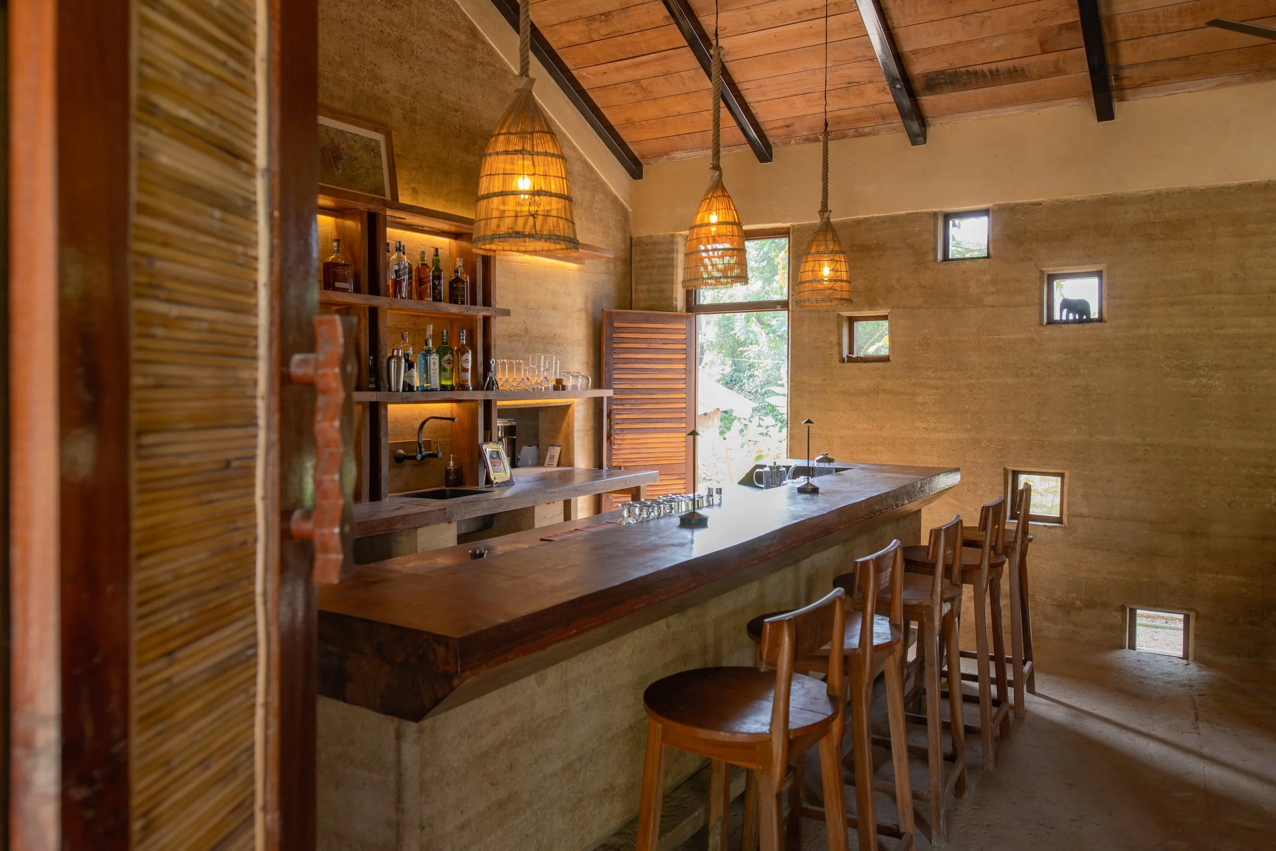 Interior of a rustic bar with wooden bar counter, hanging wicker lamps, and small square windows showing trees outside.