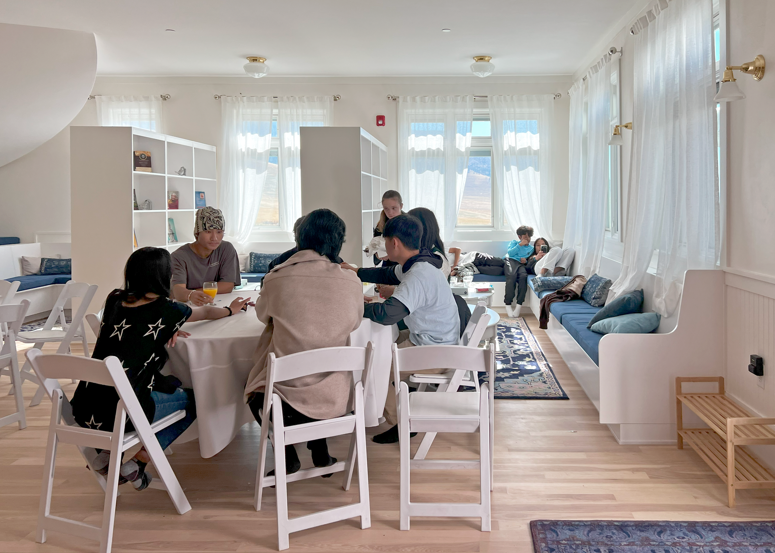 A group of people sitting around a table in a bright, airy room with large windows and white curtains.