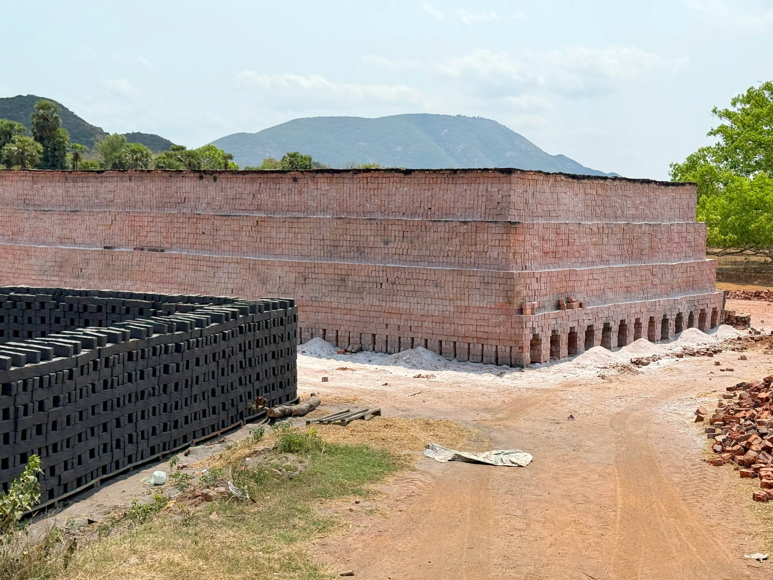 Construction site with stacked black and red bricks and a partially built brick wall, with mountains in the background and some green trees and grass around.