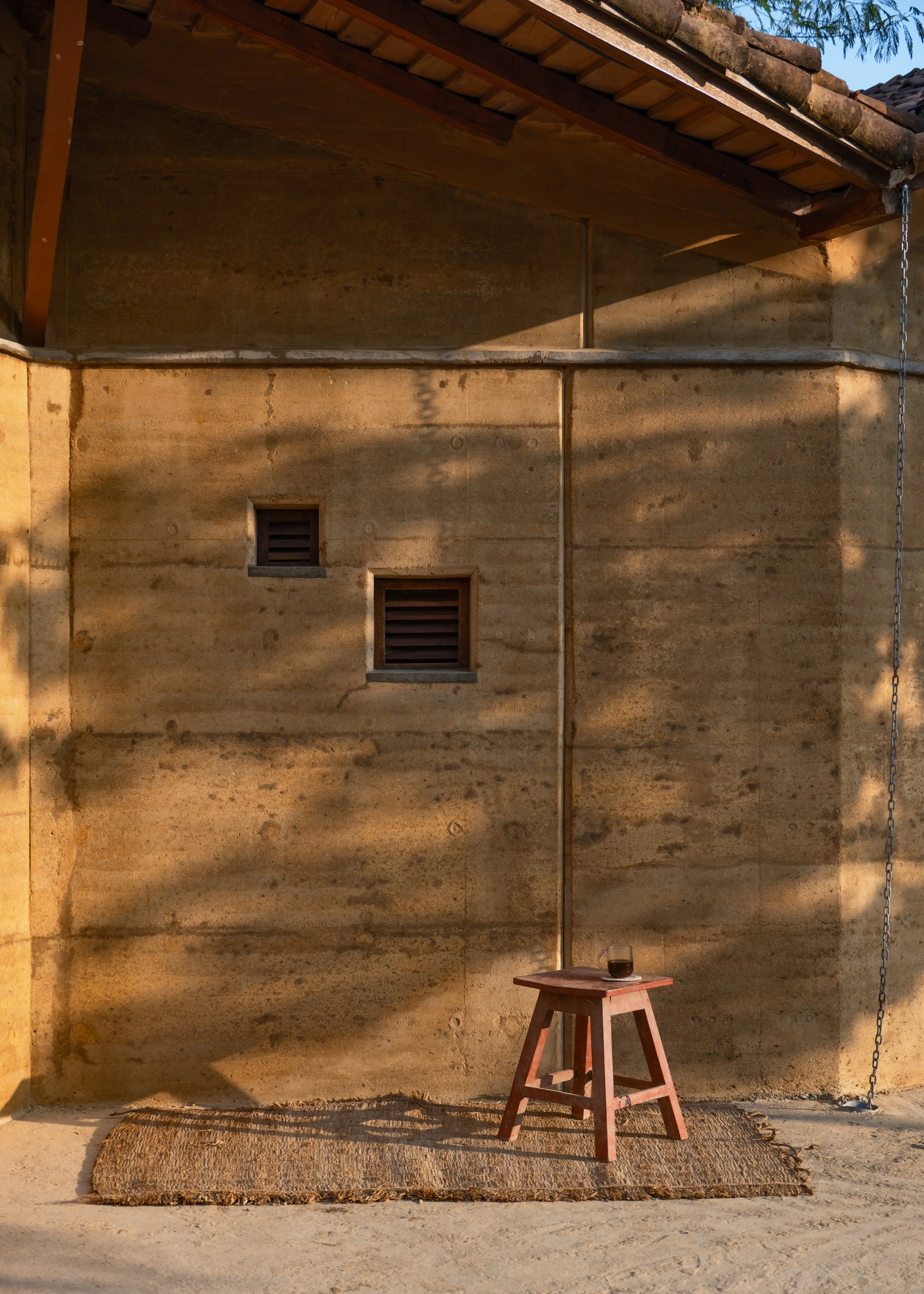 A rustic outdoor scene featuring a small wooden stool with a dark glass on top, placed on a woven rug against a weathered concrete wall with two small square ventilation openings. The scene is lit by warm sunlight, casting shadows on the wall, with p