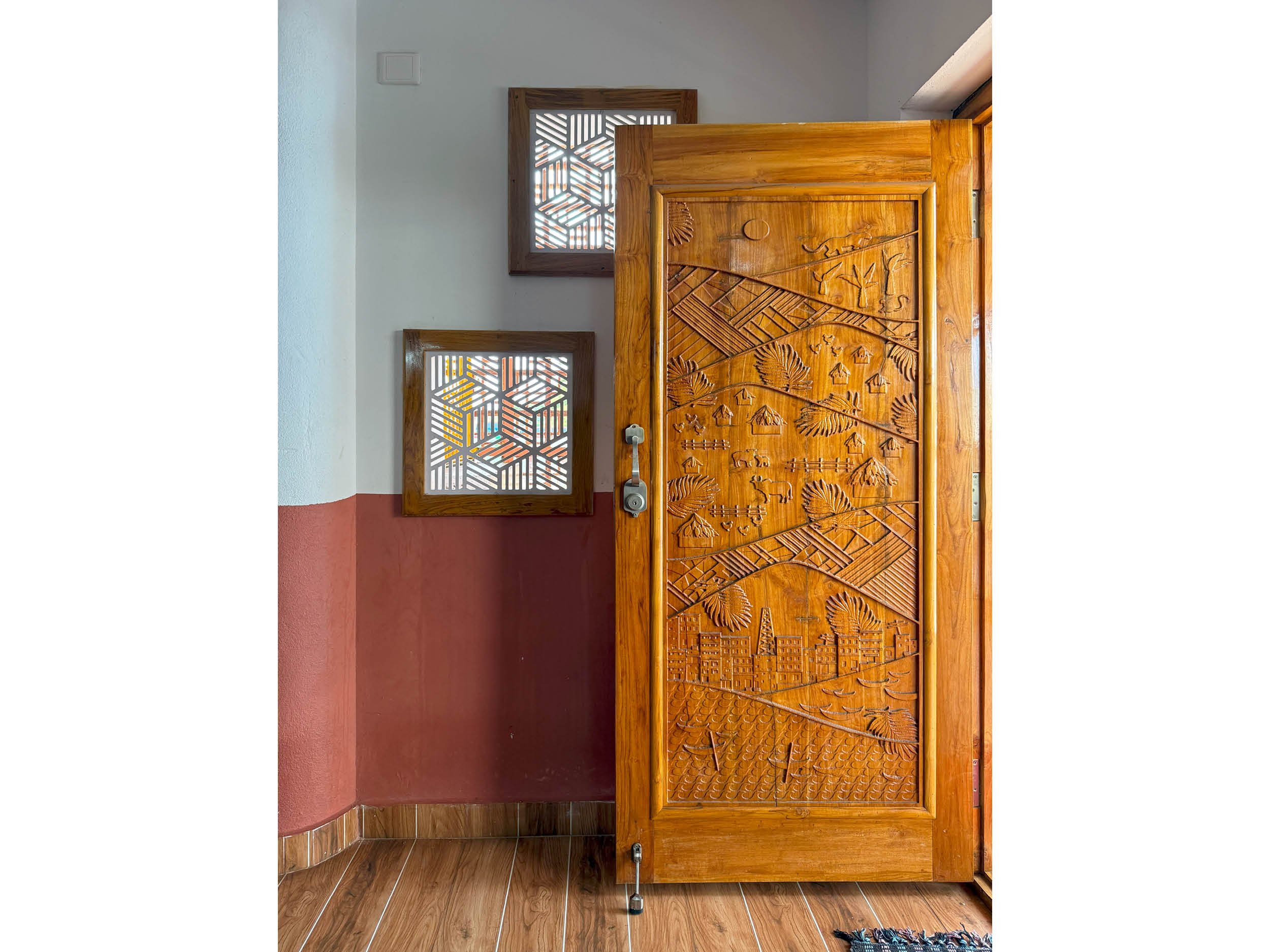 Wooden door with intricate carvings depicting scenes and nature, with a grey door handle. The room has two decorative wooden windows with geometric patterns and white walls, with a pinkish-red lower wall and brown wooden flooring.