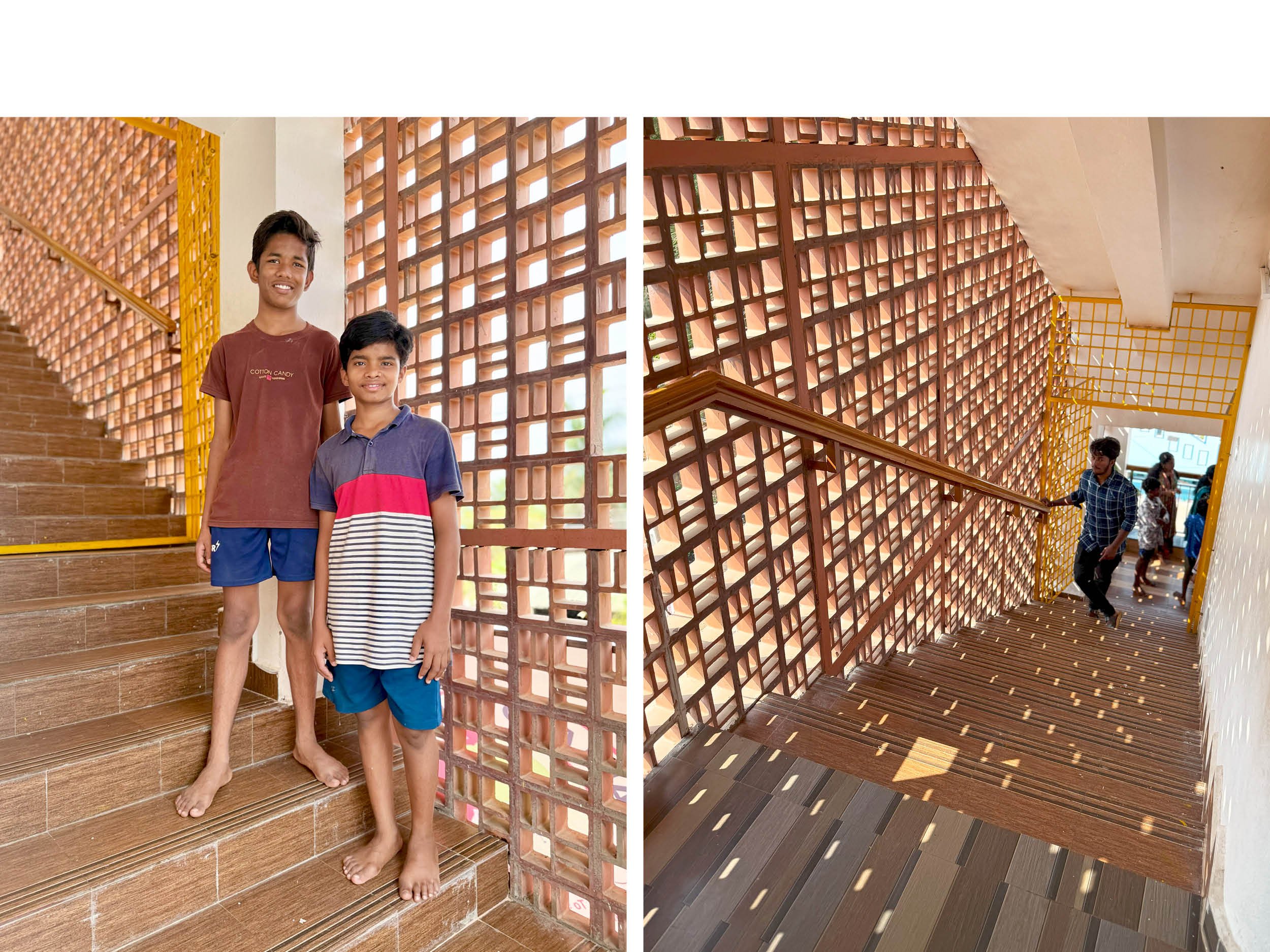 Two boys standing on stairs inside a building with brick and wooden lattice walls, one boy is wearing a brown t-shirt and the other a striped t-shirt. There are people in the background walking on the staircase.