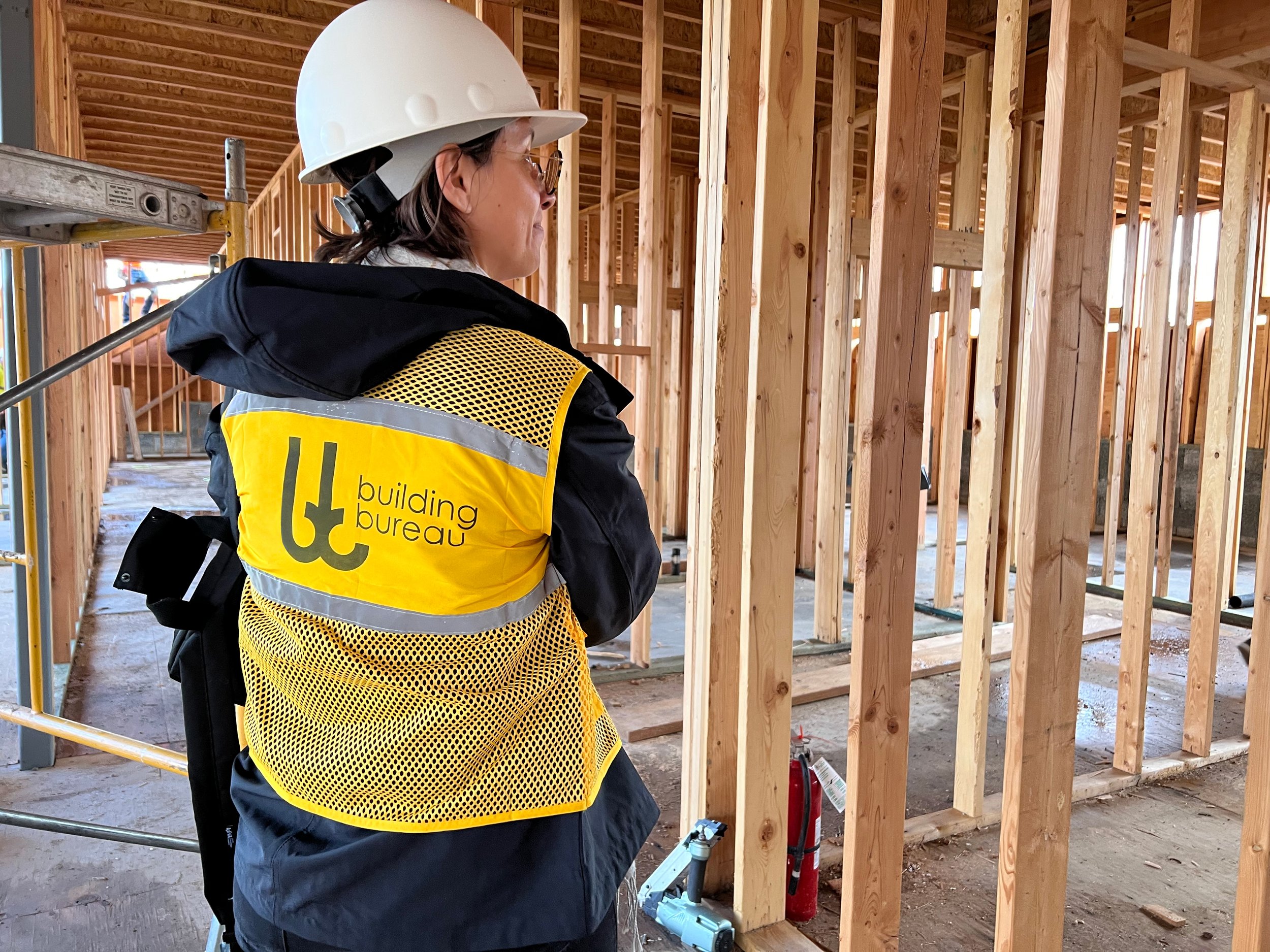 A woman wearing a white hard hat, glasses, and a yellow safety vest with black and gray details, with 'building bureau' printed on the back, inspecting a wooden framing of a building under construction.