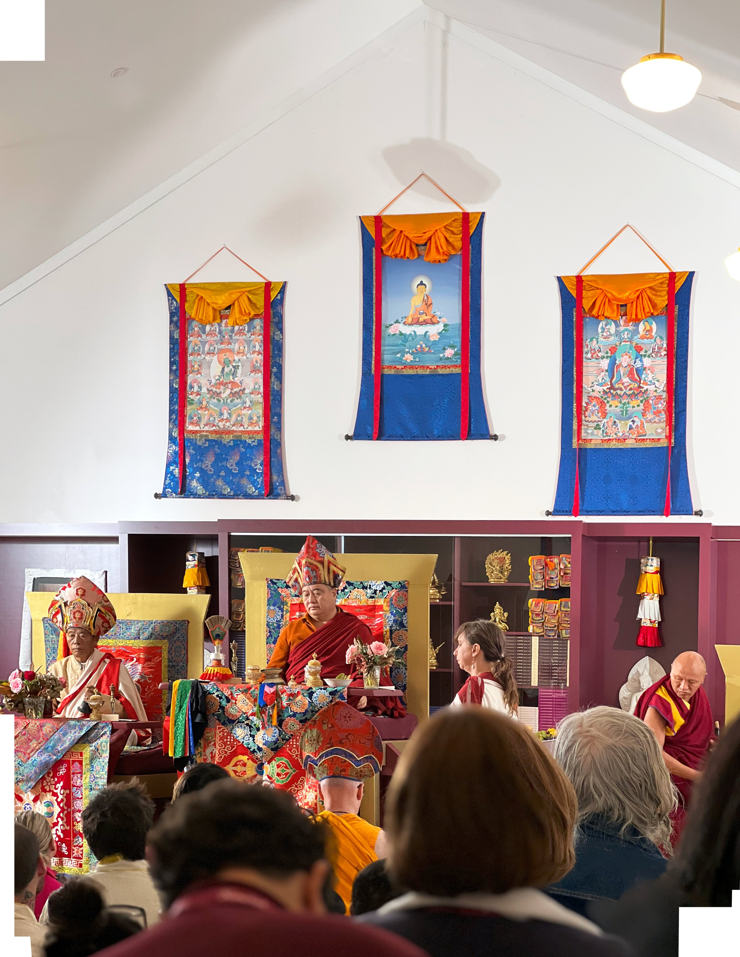 A Buddhist ceremony is taking place inside a temple. Monks and practitioners are seated in front of a shrine with colorful Buddhist artwork and statues. Three hanging banners with Theravada Buddhist images are displayed on the wall.