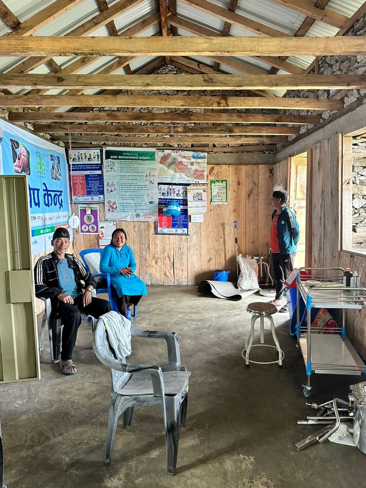 Interior of the Ekuwa Health Post with wooden walls and ceiling, showing two seated people and medical equipment and posters on the wall.