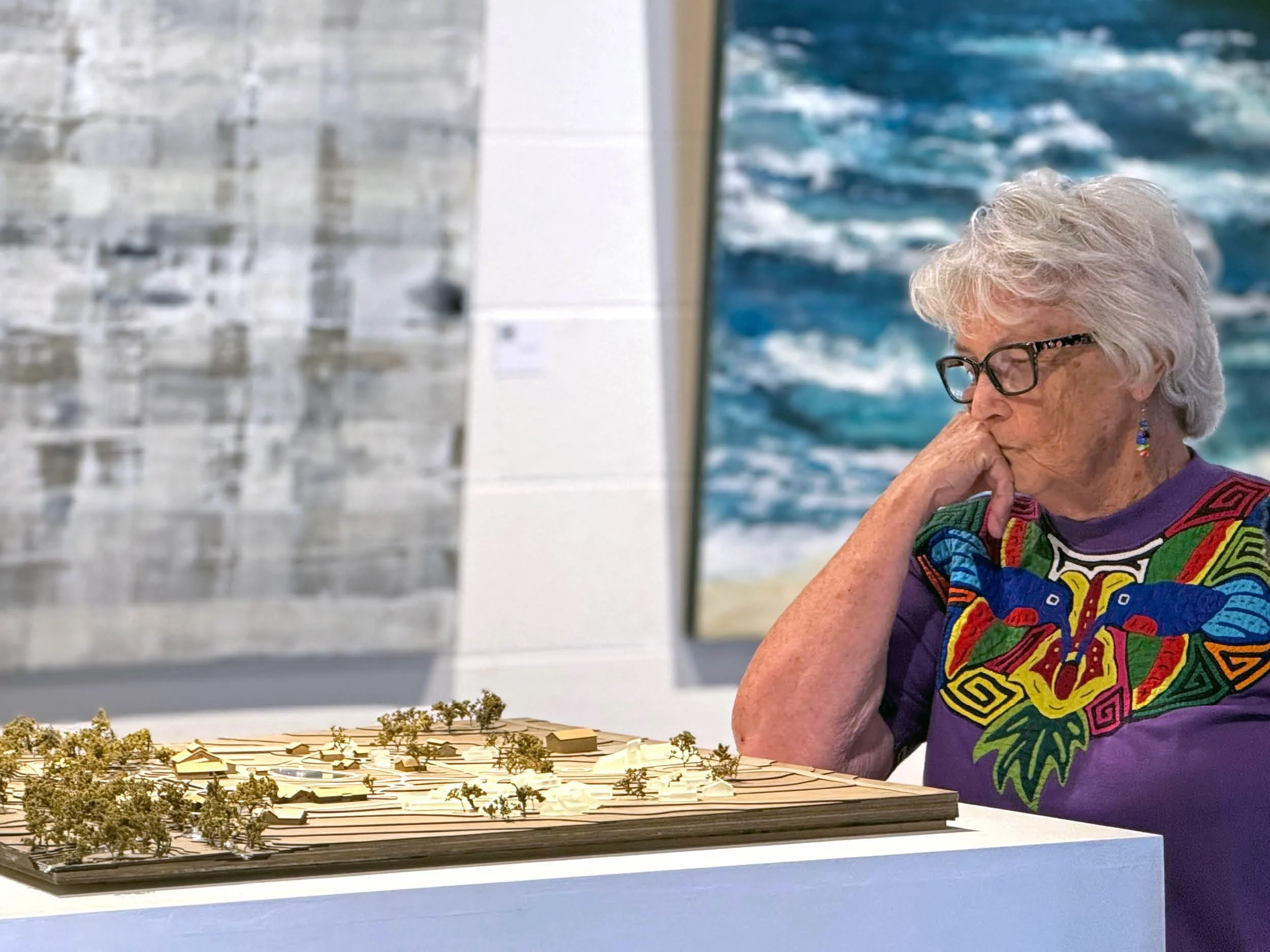 An elderly woman with white hair, glasses, colorful earrings, and a vibrant patterned shirt examines the model of the Lighthouse Ranch by Building Bureau.