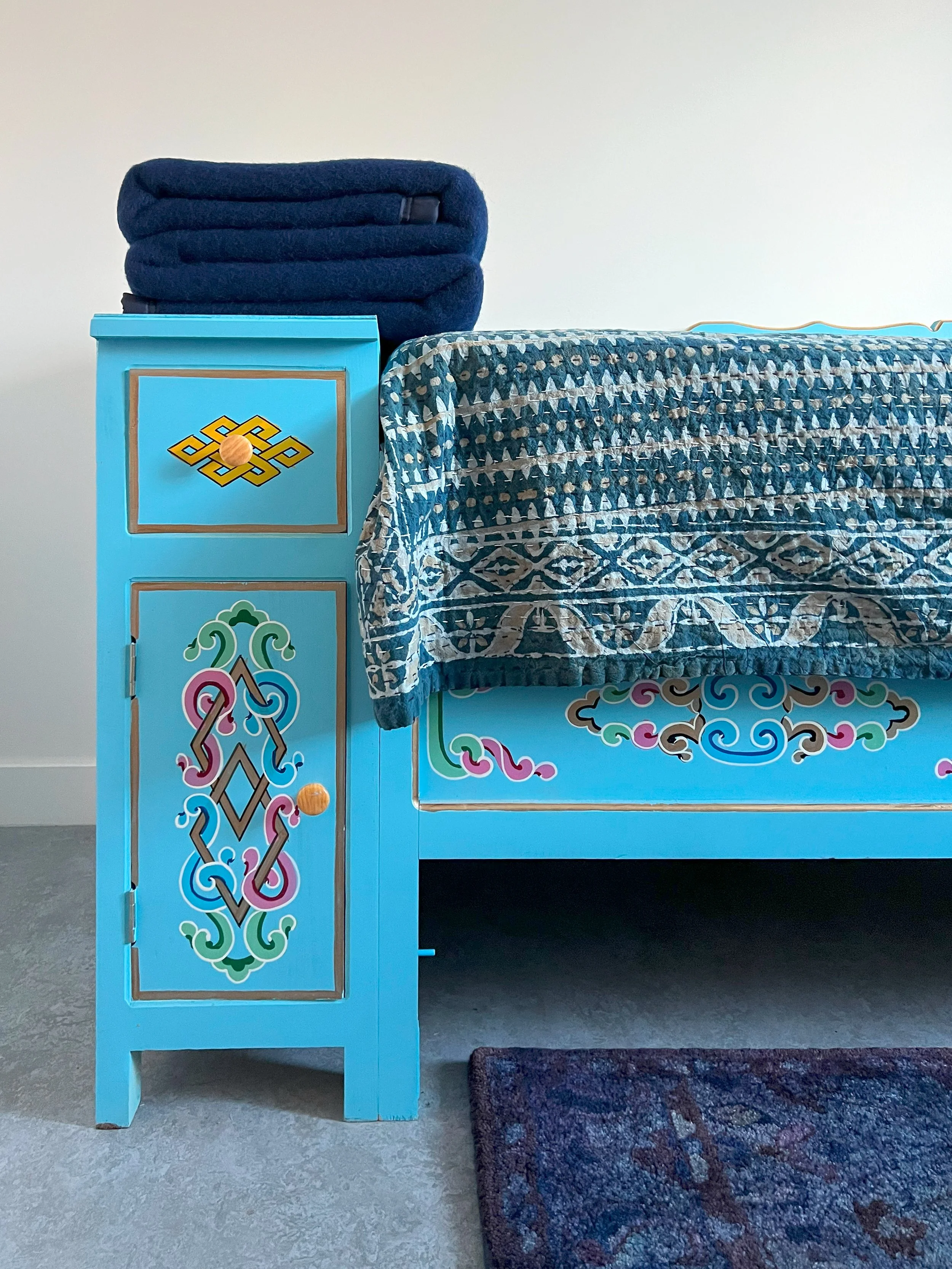 A blue piece of furniture with colorful decorative patterns on the sides, next to a table with a patterned cloth and a pile of folded blue blankets on top.