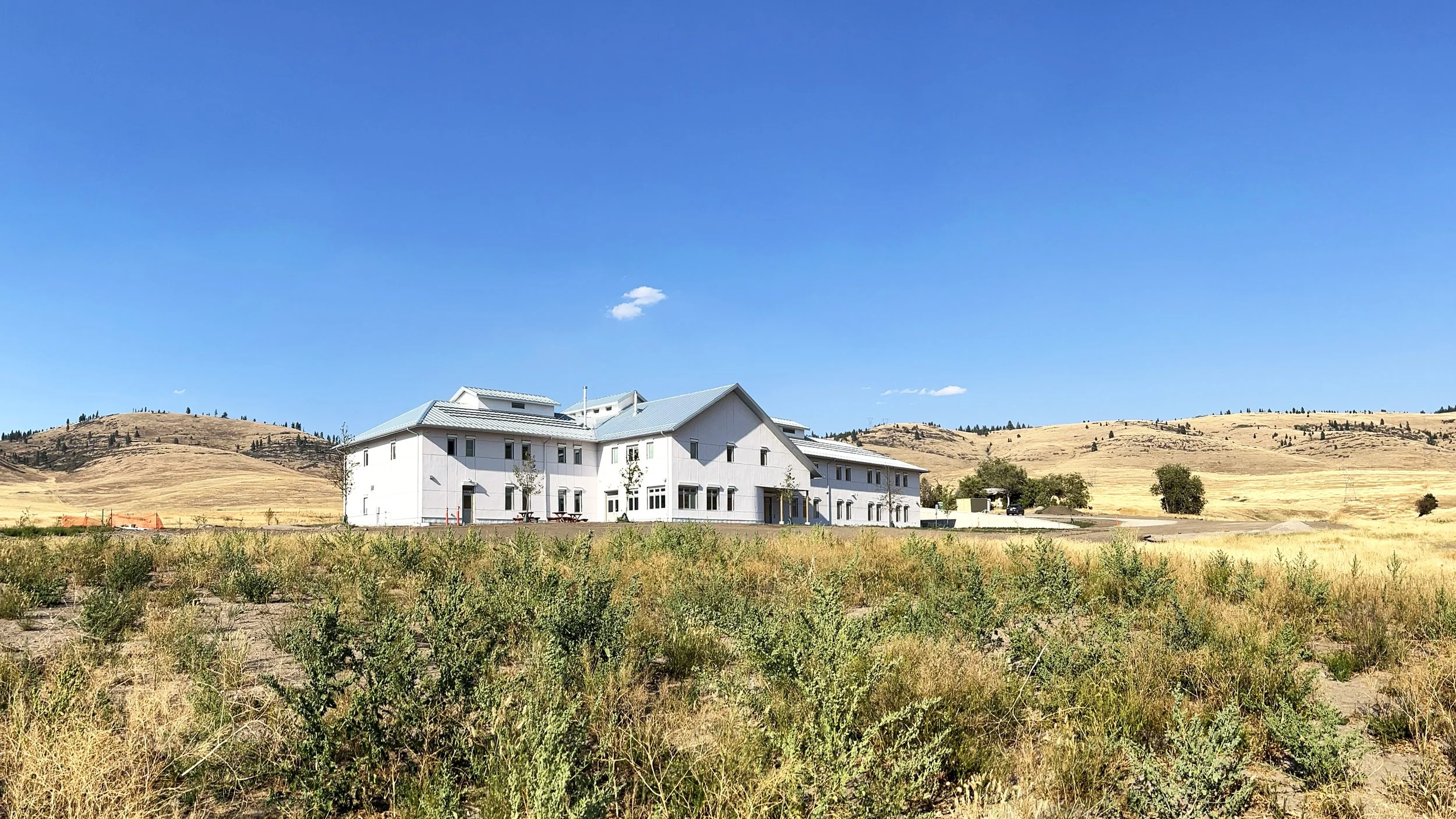 White multi-story building with a gray metal roof in a rural landscape with rolling hills and sparse trees under a clear blue sky.