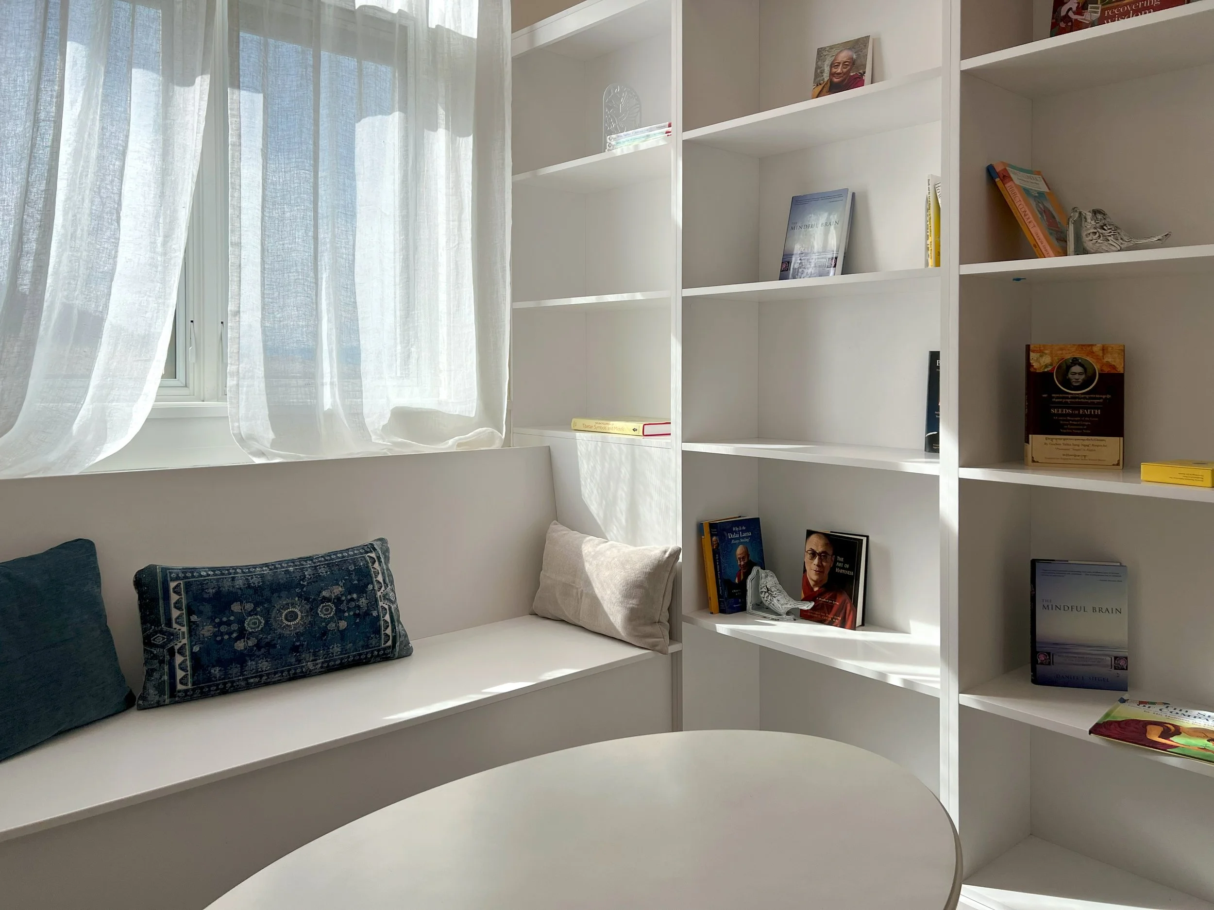 Sunlit corner with white bookshelf filled with books and decorative objects, next to a window with sheer white curtains, and a white cushioned window seat with patterned pillows.