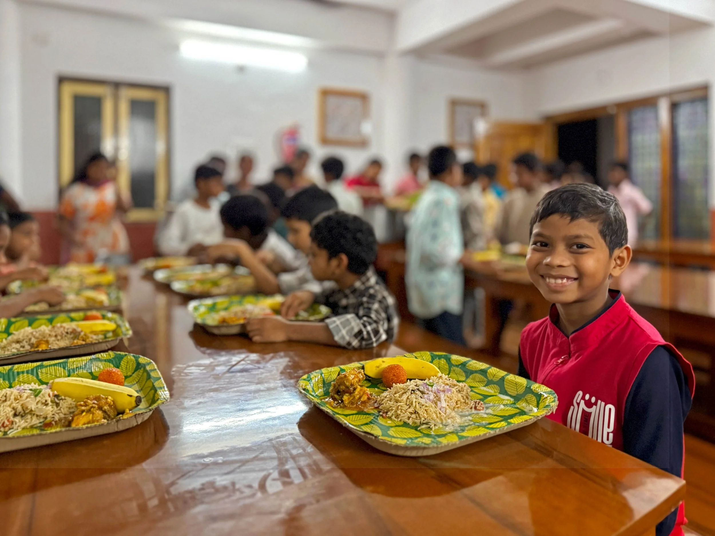 A smiling young boy in a red shirt is seated at a table with plates of food, with more children and adults in the background in a brightly lit indoor setting, possibly a classroom or community hall.