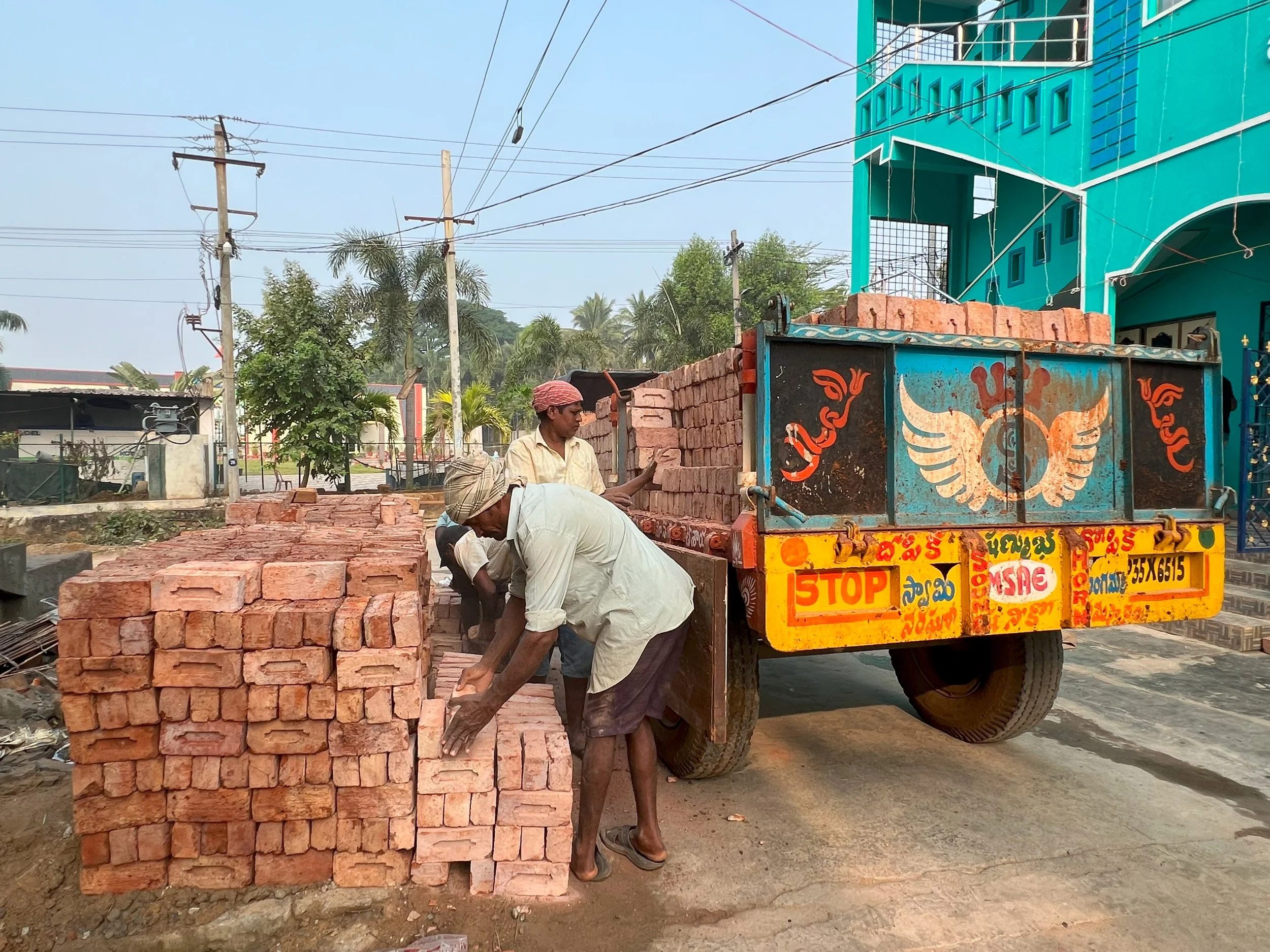 Men working with bricks around a truck filled with bricks, in front of a bright blue building with greenery and utility poles in the background.