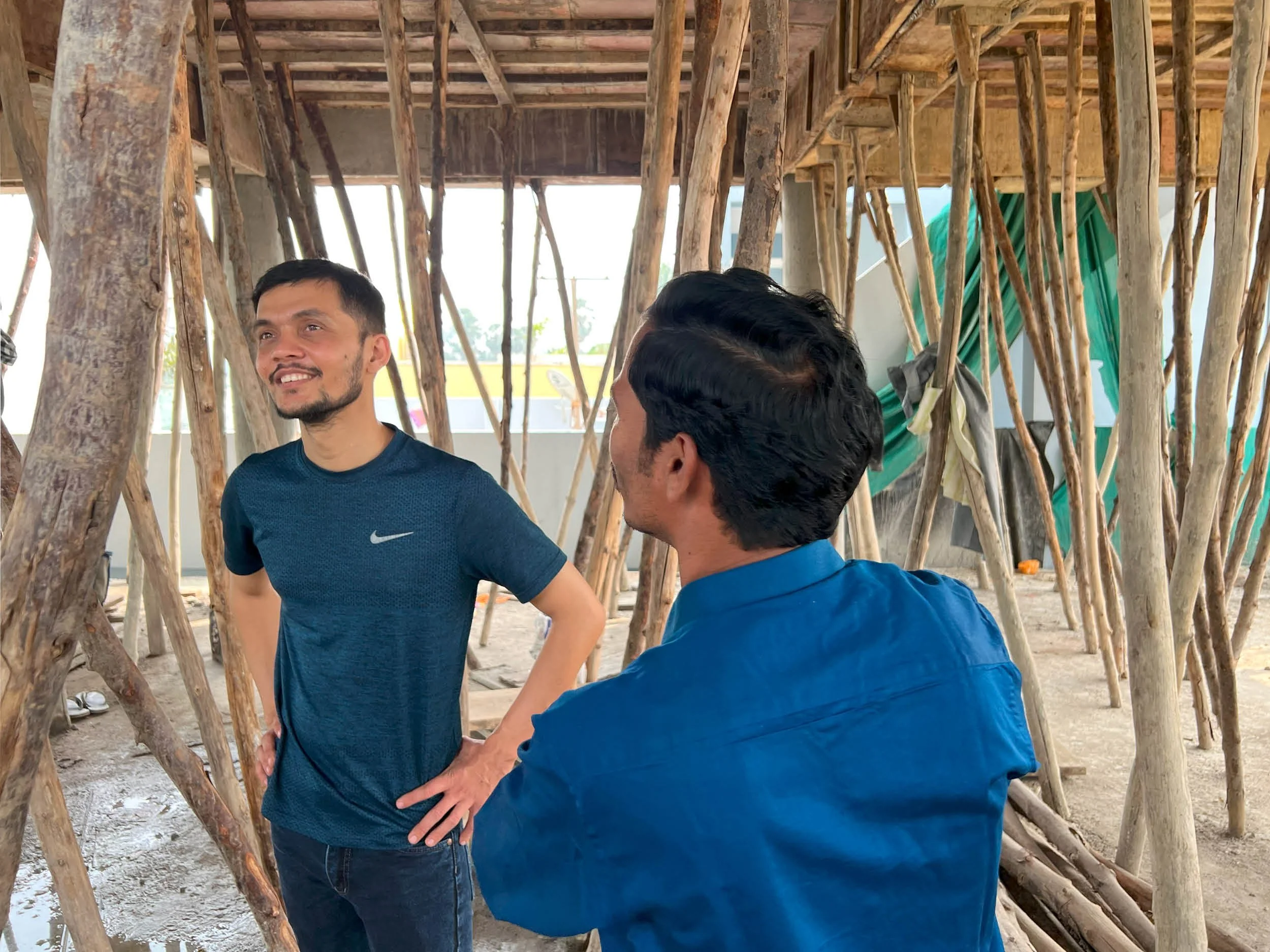 Two men talking inside a wooden structure made of tree branches with a partially constructed roof.