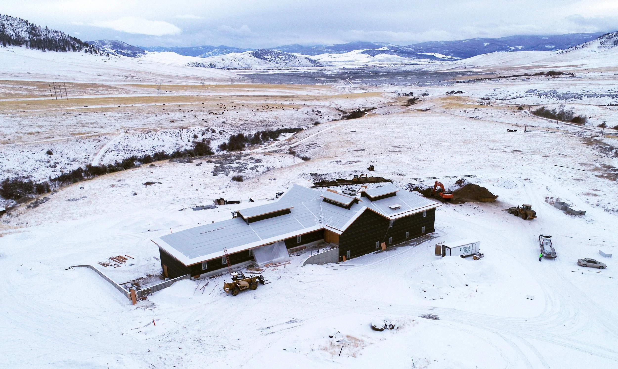A snow-covered landscape with rolling hills in the background. In the foreground, there is a building under construction with a metal roof, construction vehicles, and equipment. The scenery includes scattered bushes, a few trees, and a construction s