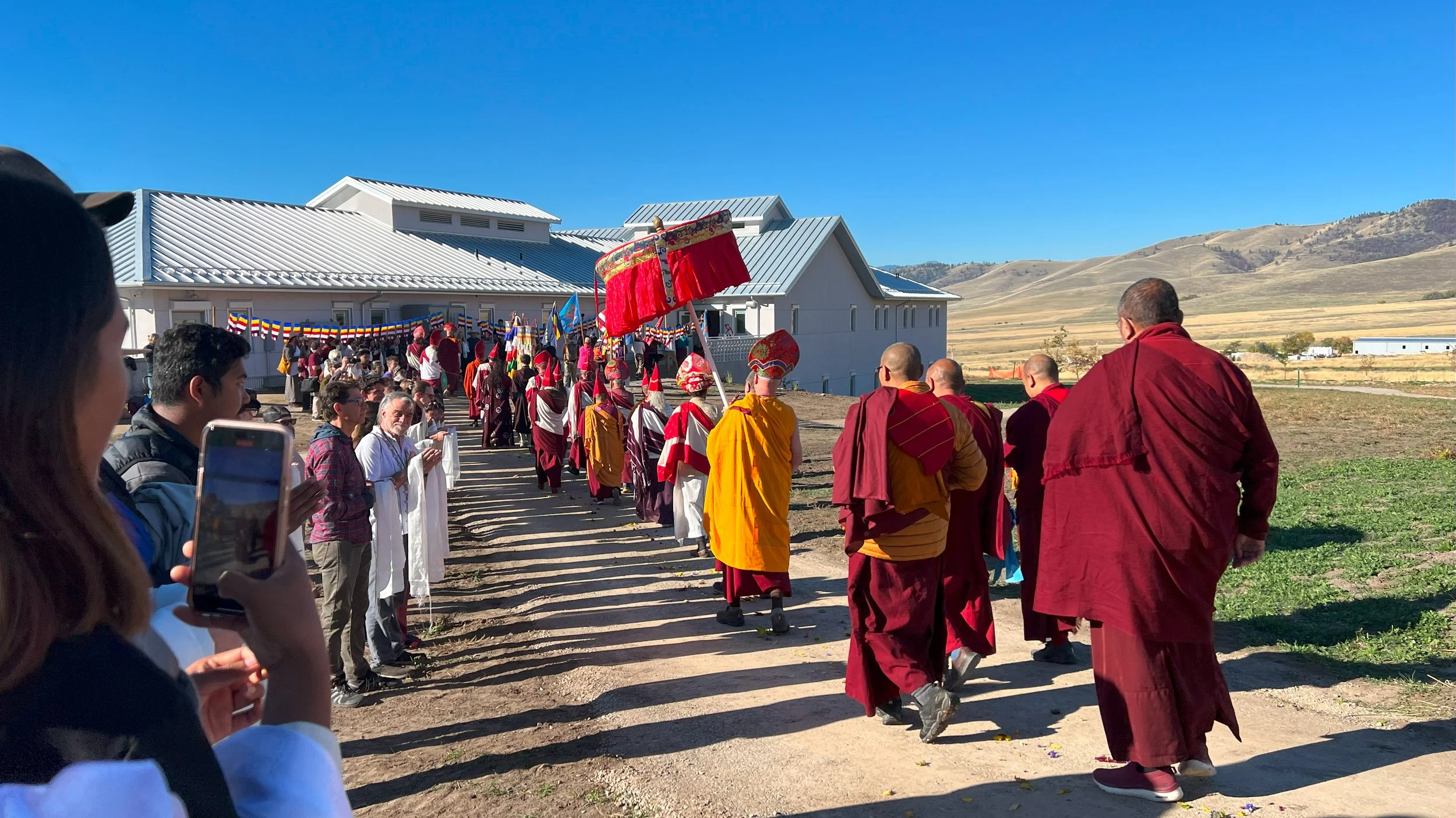 A group of monks and laypeople participating in a cultural or religious procession outdoors, with a large building and rolling hills in the background on a clear day.