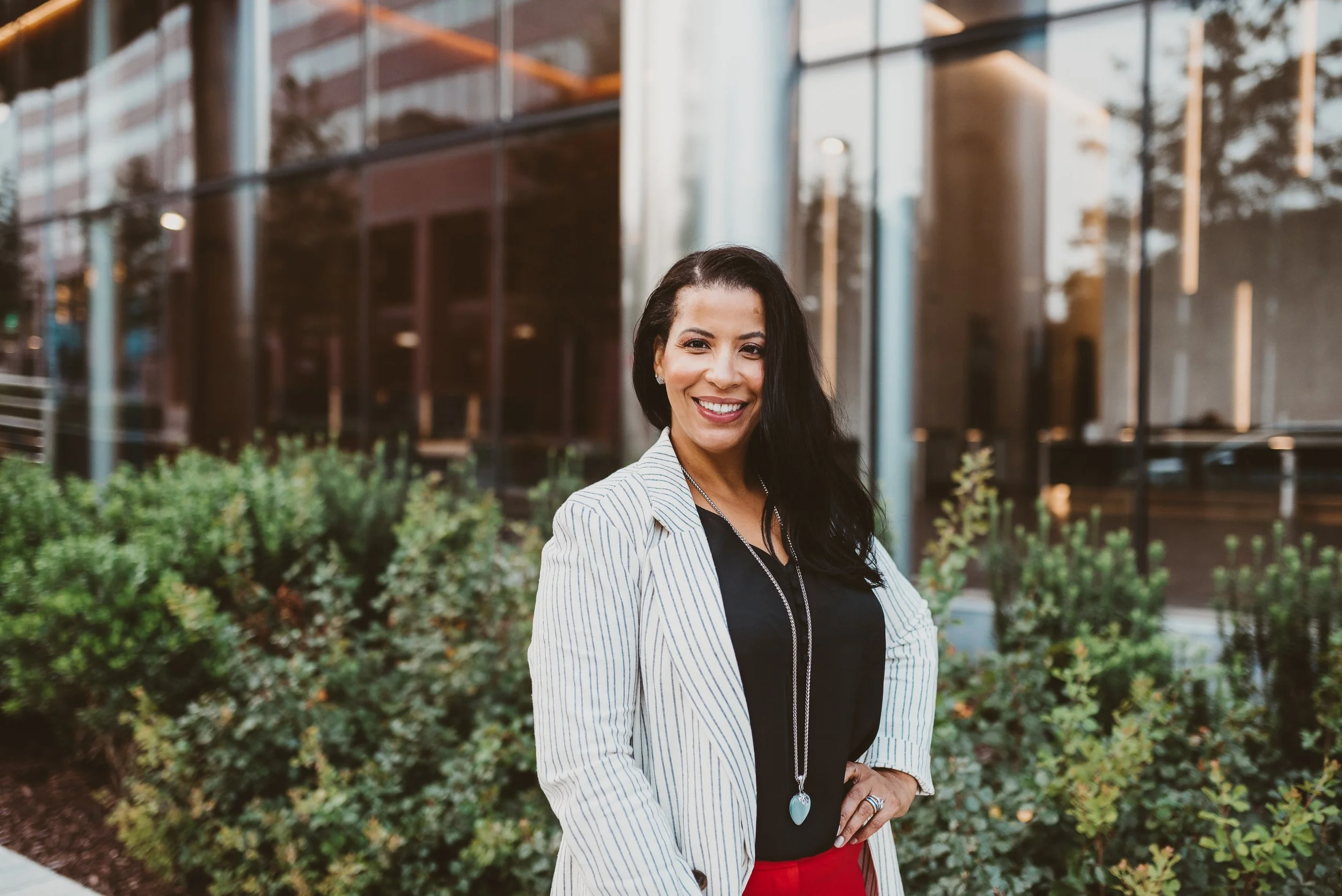 A smiling woman with long dark hair standing outdoors near a modern glass building with greenery in the foreground.
