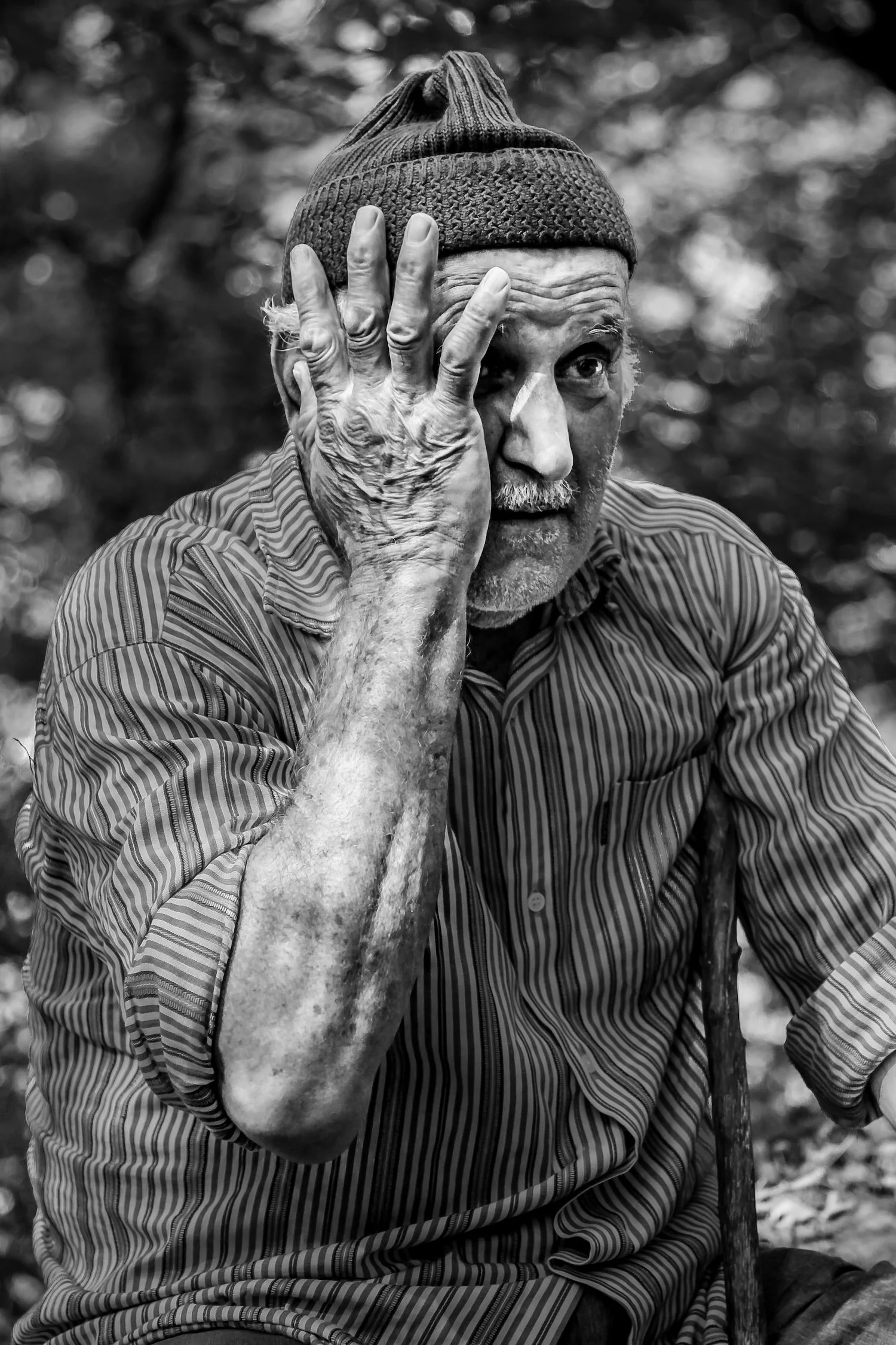 An elderly man with a beanie and striped shirt, resting his head on his hand with a road and forest in the background