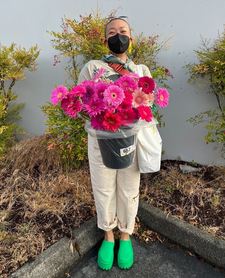 Monyee, a nonbinary person, standing in front of a gray wall with outdoor plants. They are wearing a beige outfit with lime green shoes and a forest green scarf around their neck. They are holding a bucket full of magenta daisies.