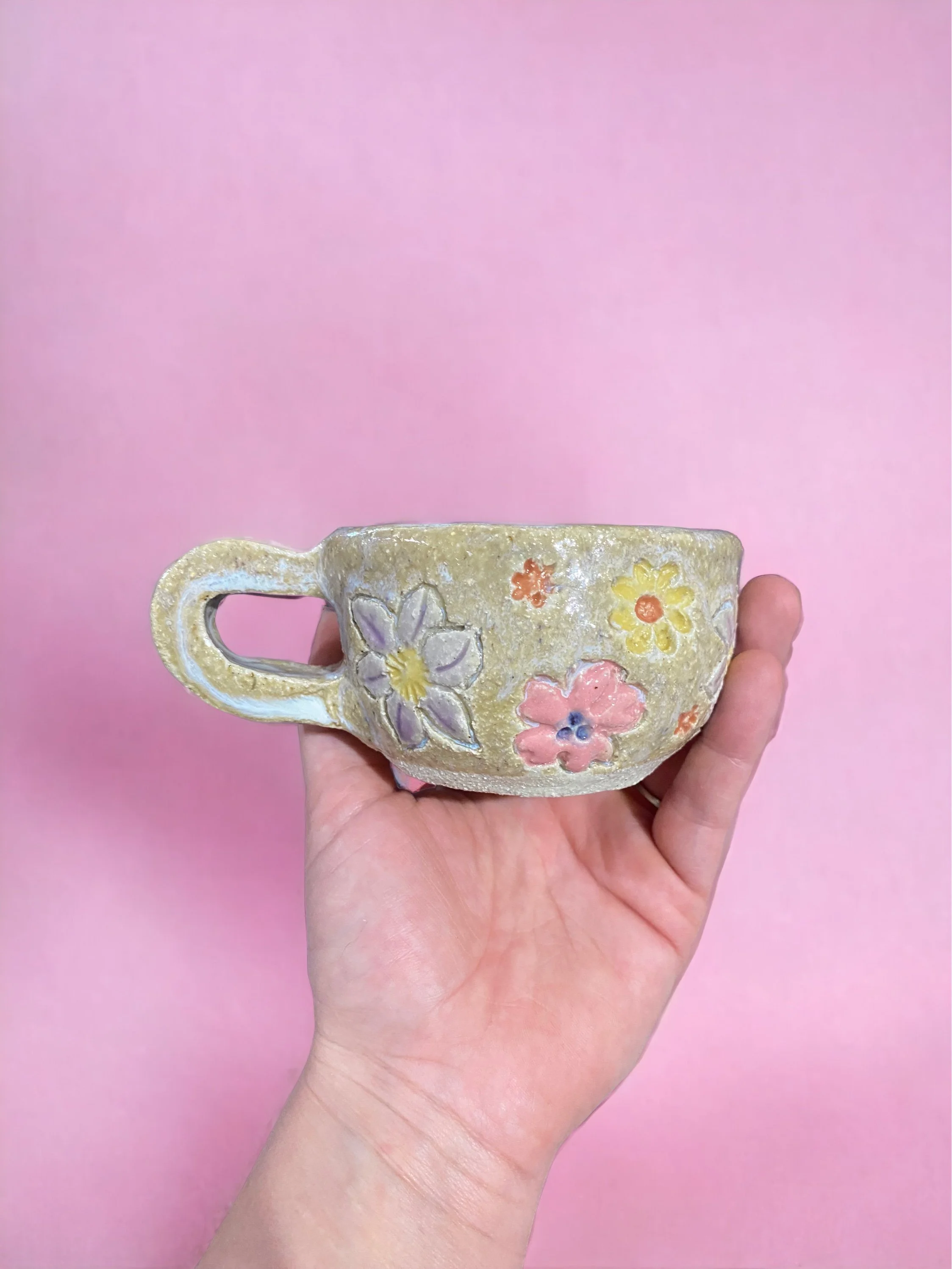 Hand holding a ceramic mug with pastel-colored floral designs against a pink background.