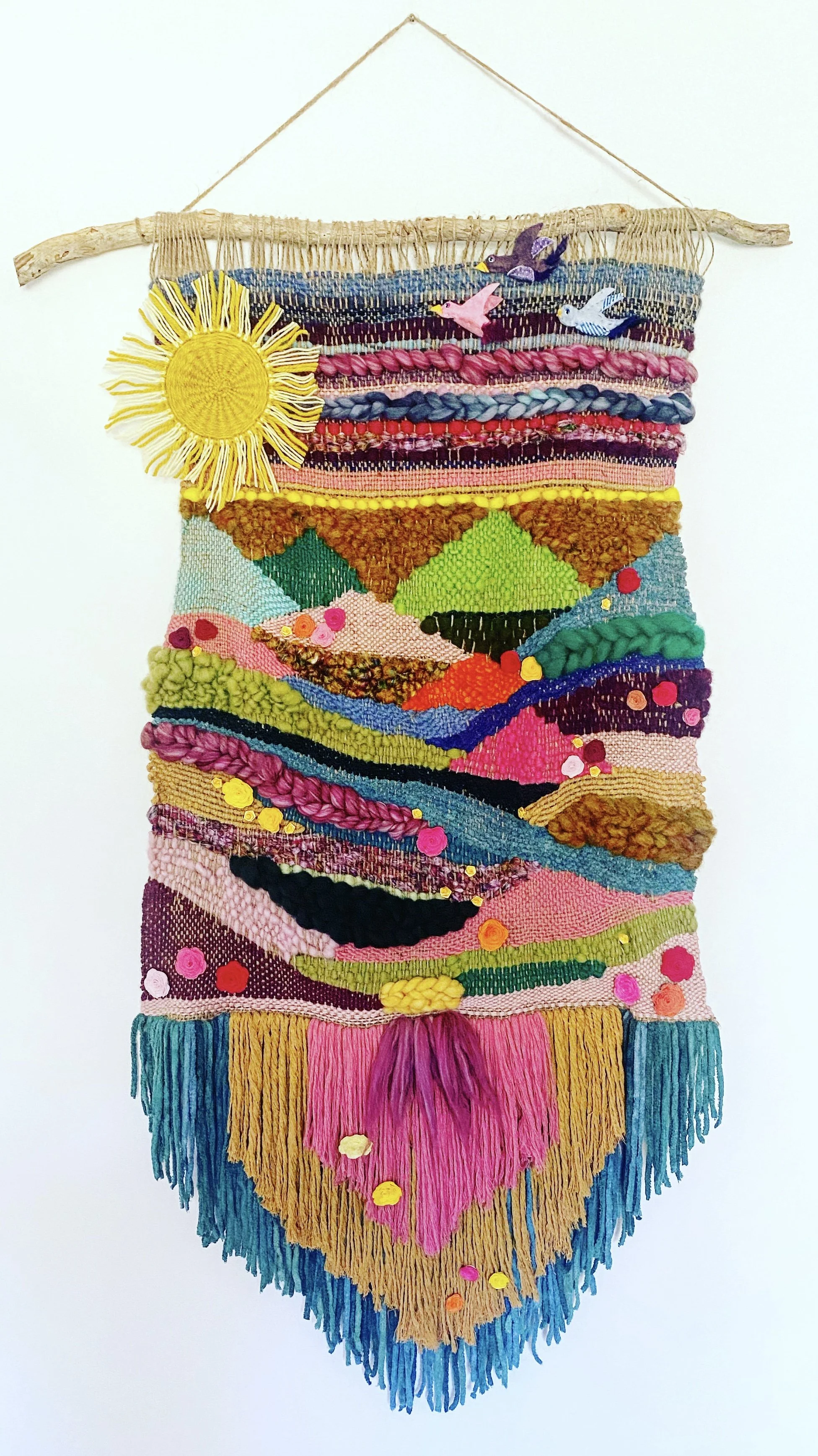 Colorful woven wall hanging with a yellow sun, pink and purple birds, and a landscape scene with mountains, rivers, and fields, on a white wall.