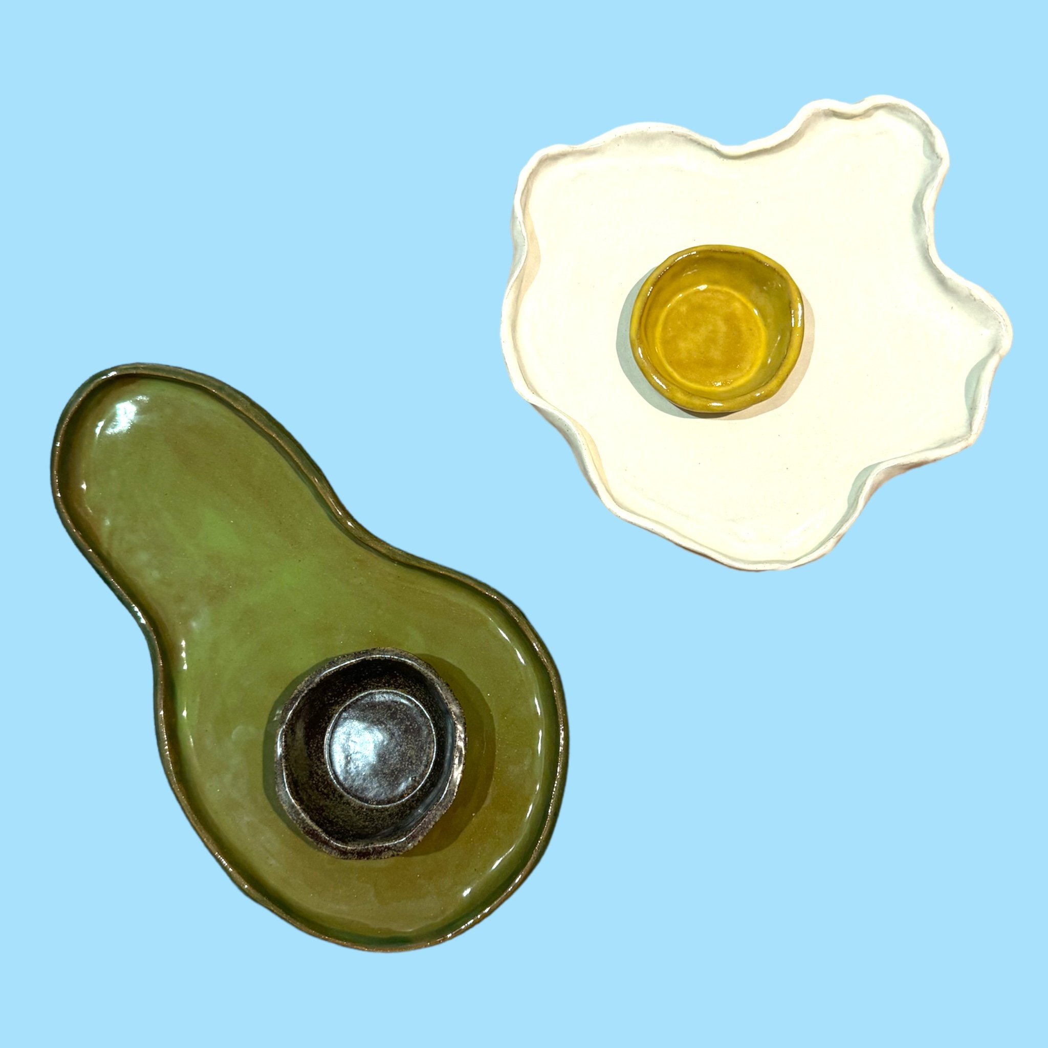 Two decorative ceramic dishes shaped like an avocado and a fried egg on a blue background.