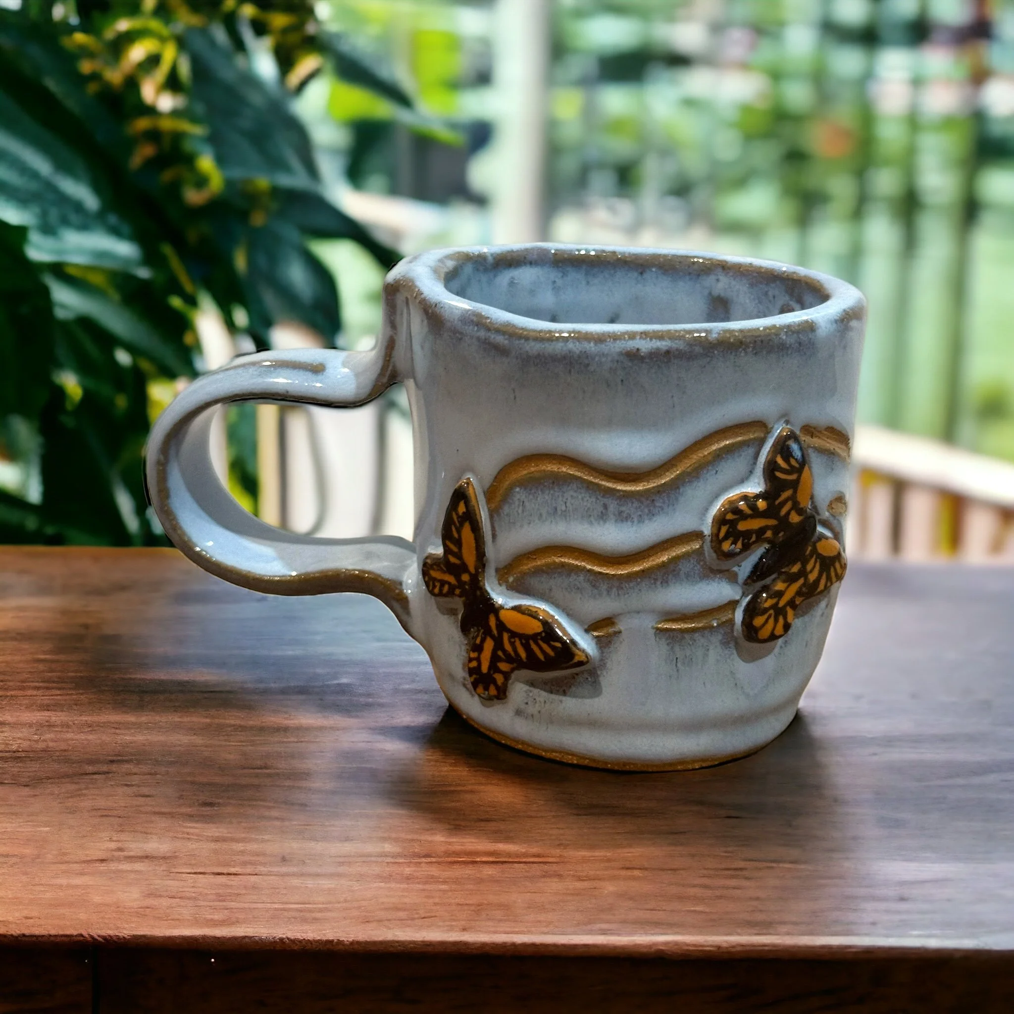 A white ceramic mug with gold and black butterfly designs and wavy gold lines, placed on a wooden surface with a green outdoor background.