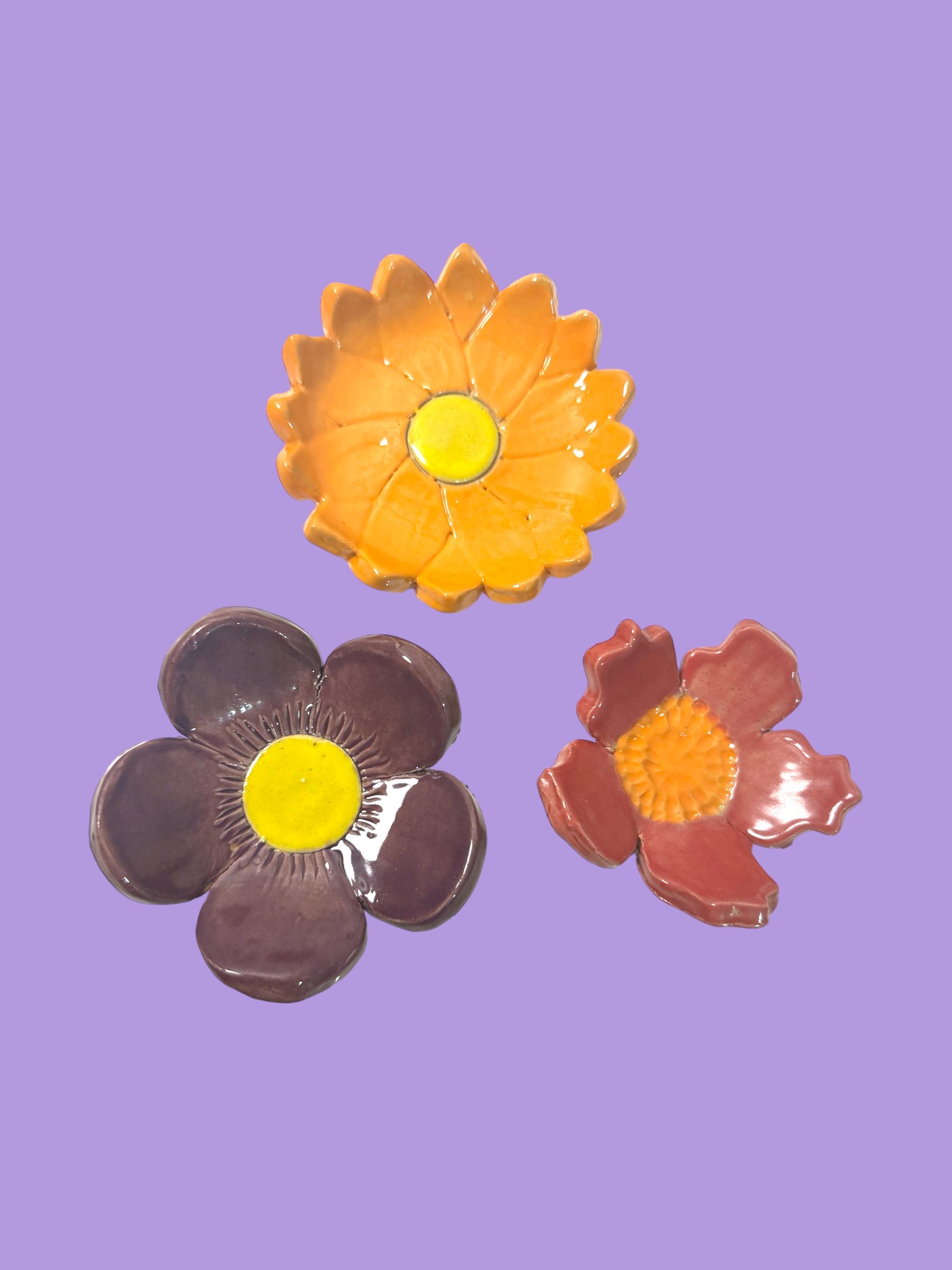 Three ceramic flower decorations, one yellow, one purple, and one pink, on a purple background.