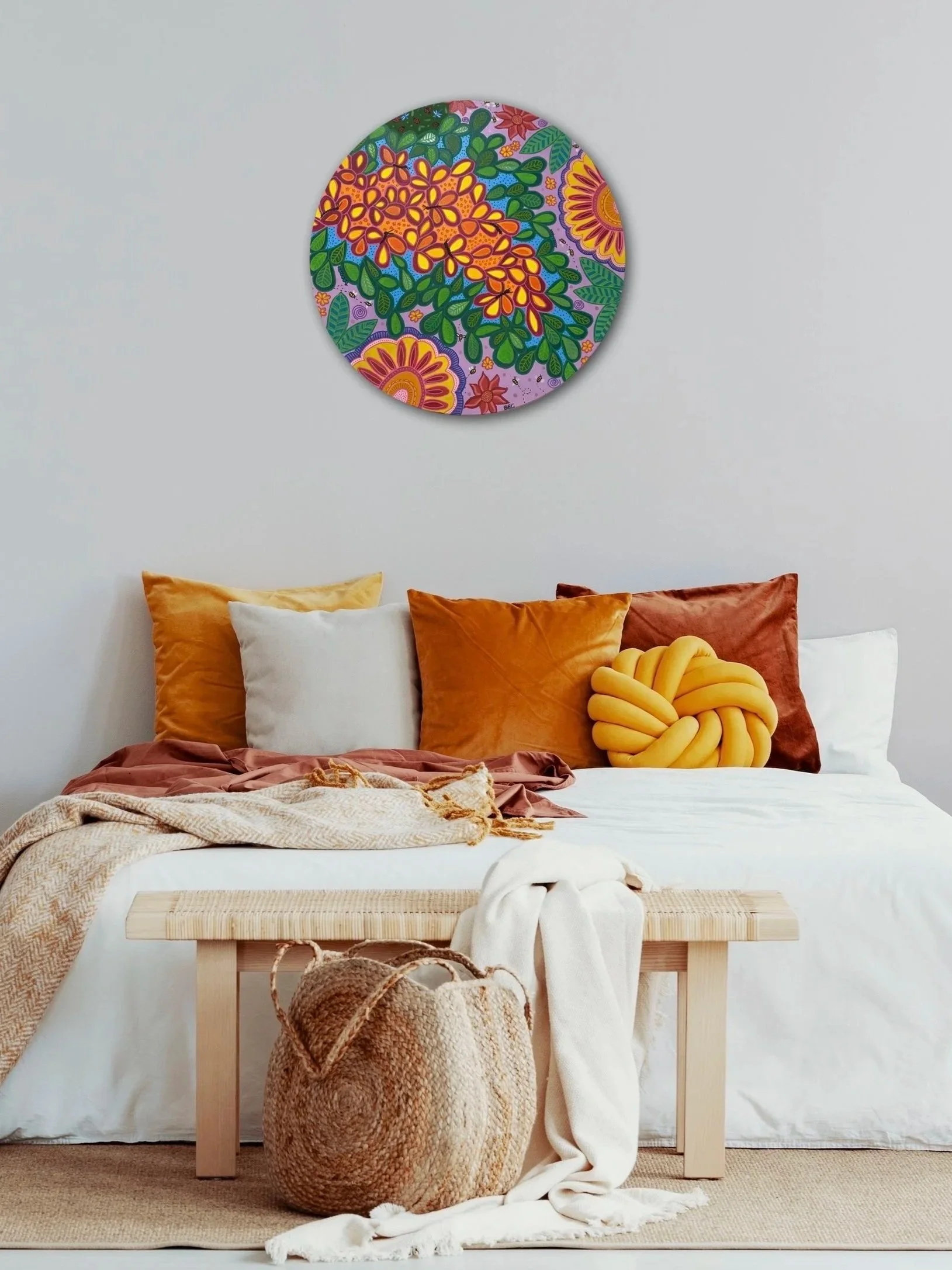 A bedroom with a white bed, surrounded by orange, white, and rust-colored pillows. A beige throw blanket and a cream-colored fabric are on the bed. There's a woven basket with a white fabric and a wooden bench at the foot of the bed. A colorful round