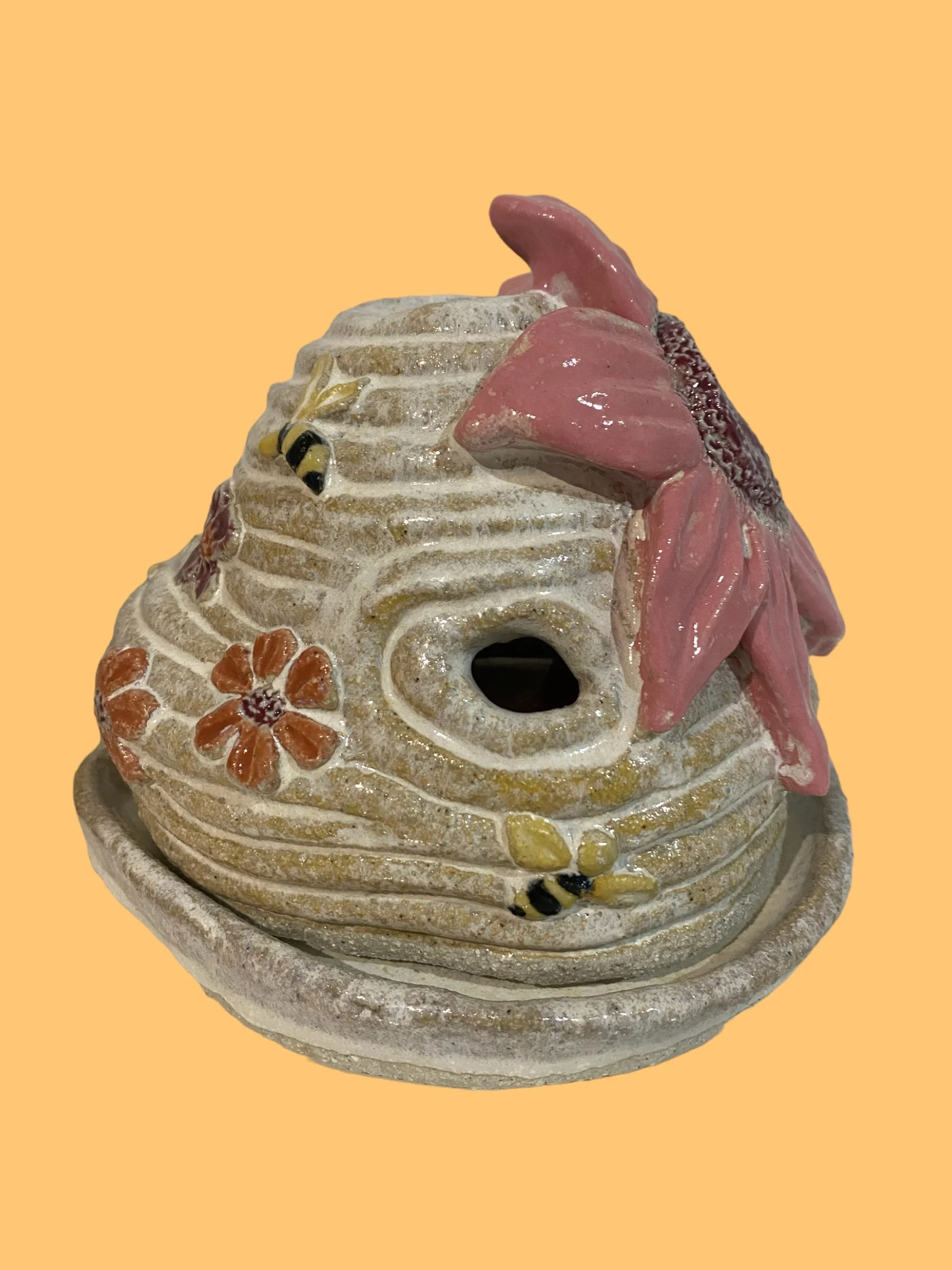 Decorative ceramic birdhouse with pink flowers, bees, and a bee on the ground, on a circular base, against a beige background.