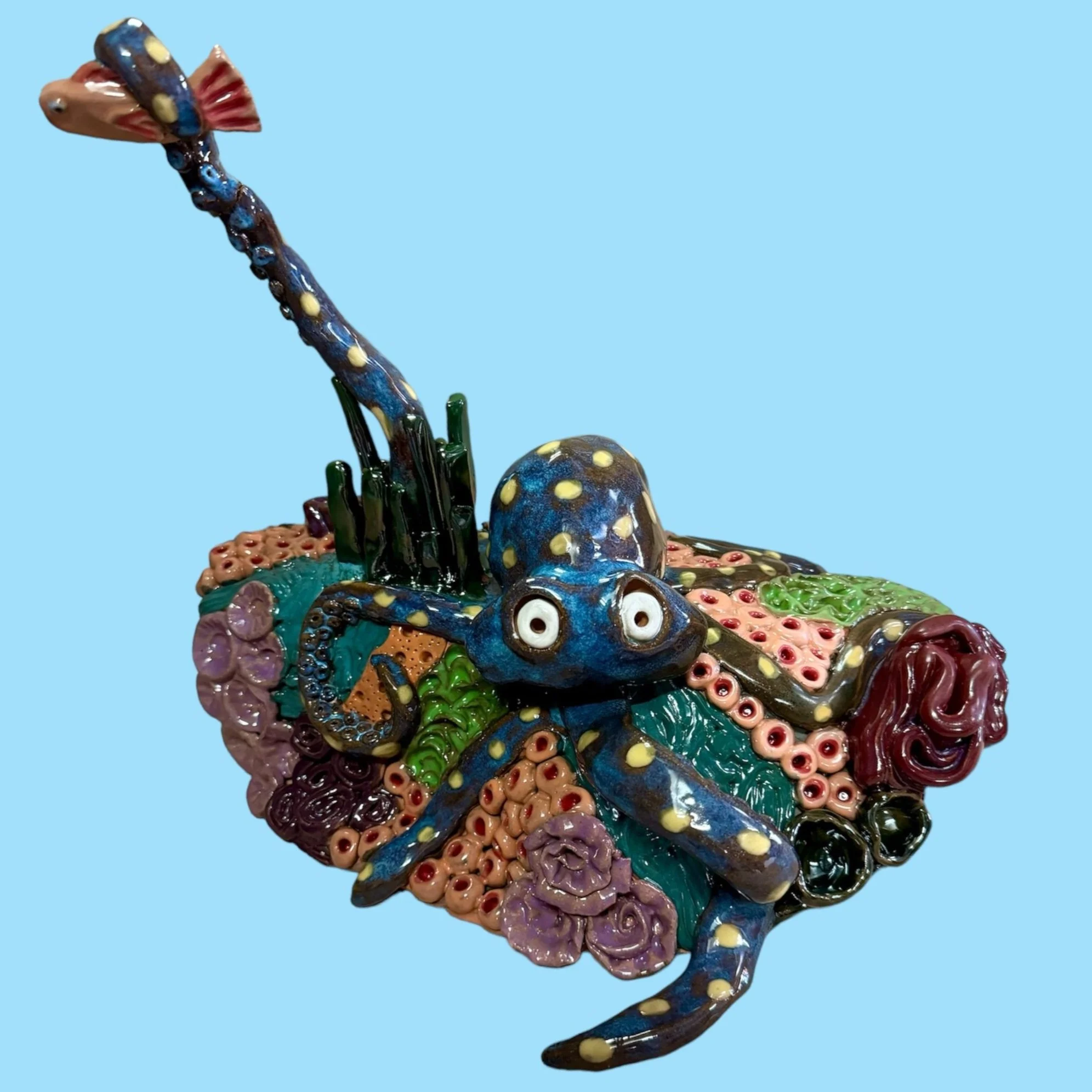 Colorful ceramic sculpture of a blue octopus with yellow spots, surrounded by various other sea creatures and coral-like formations on a light blue background.