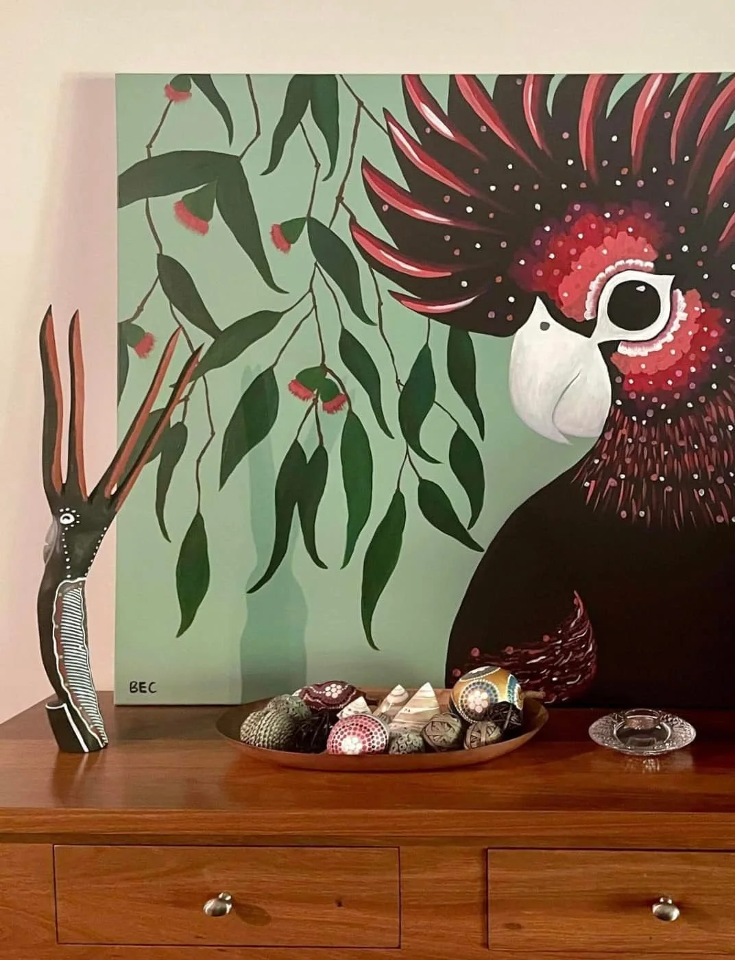 A painting of a red and black cockatoo with a white beak sits on a wooden surface, with a green leafy background. There is a tall, painted wooden sculpture of a bird on the left and a tray of decorative balls, shells, and stones in front. A small gla