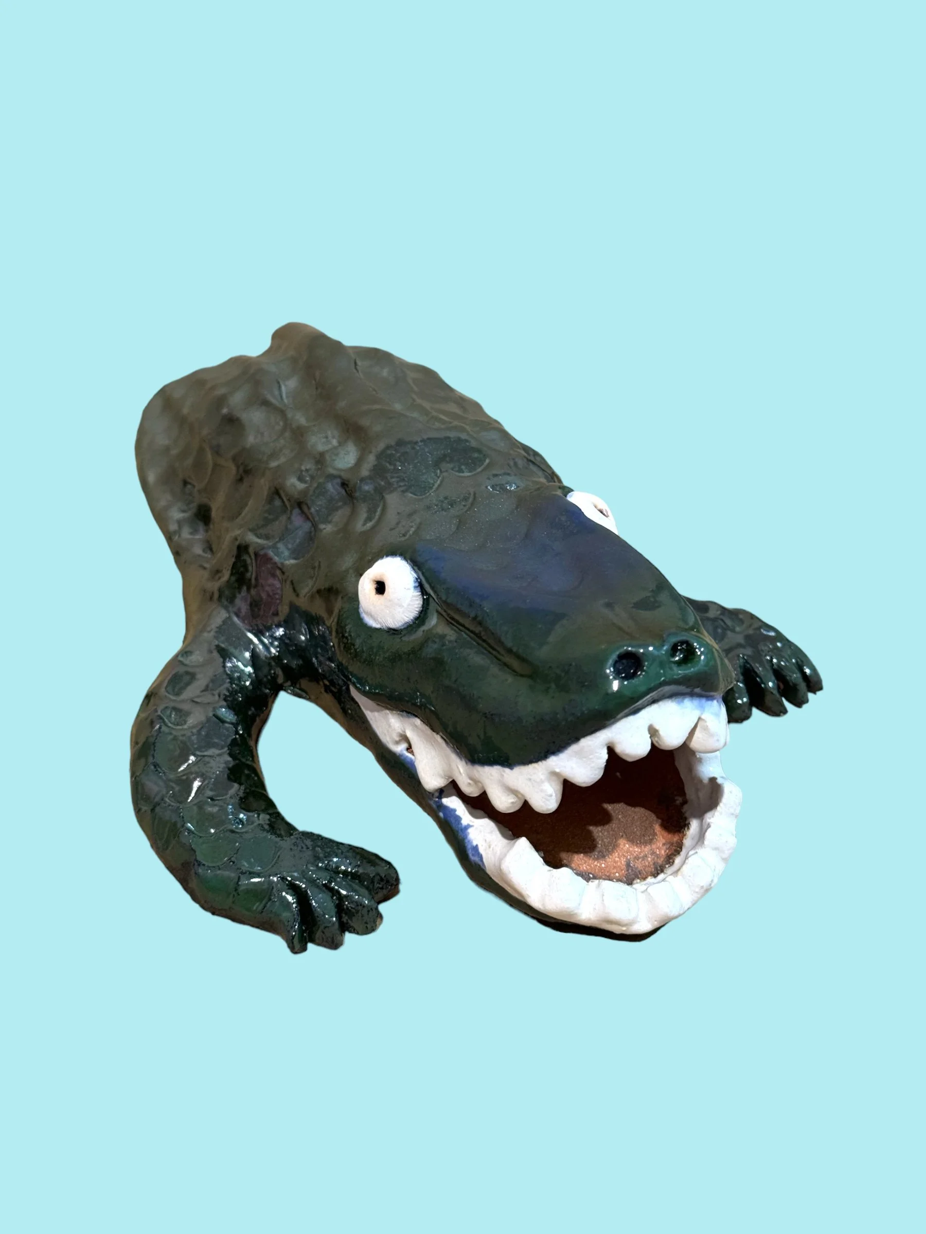 A ceramic sculpture of a crocodile with an open mouth, detailed teeth, and googly eyes, set against a light blue background.